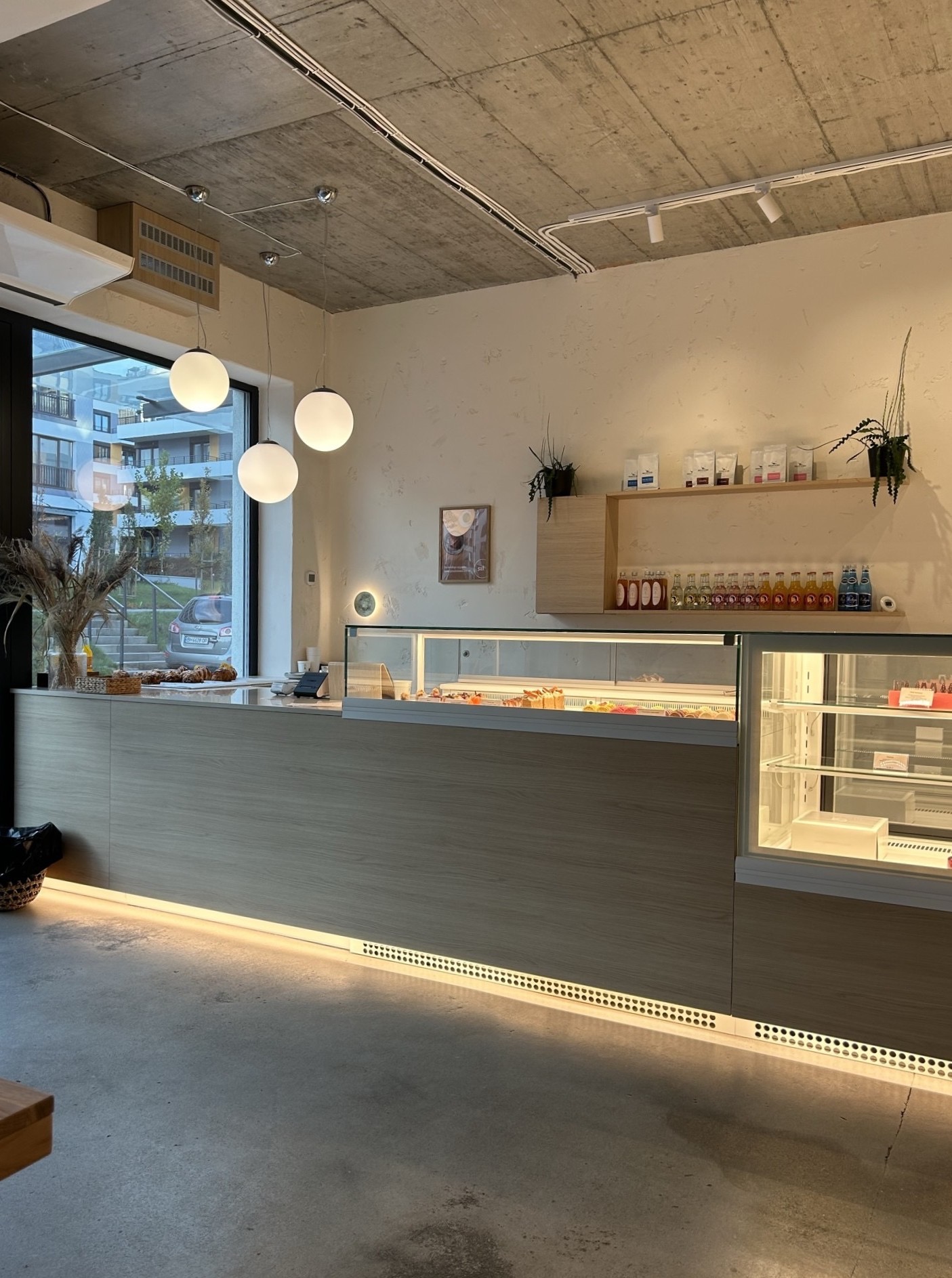 Nowoczesne wnętrze kawiarni z oświetlonym ladą, przeszkloną witryną z deserami i minimalistycznymi półkami na ścianie z napojami i kawami.