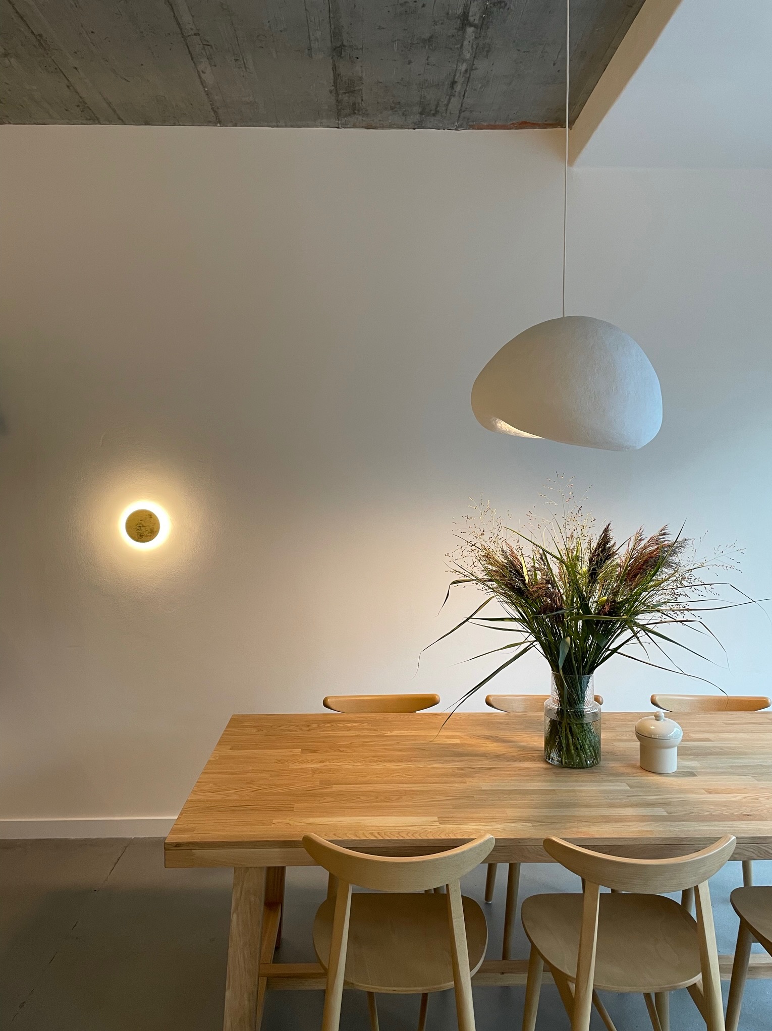 Jasna jadalnia: drewniany stół, krzesła, lampa w kształcie chmury. Na stole wazon z trawami i ceramiczny pojemnik. Minimalistyczny design, surowy betonowy sufit.