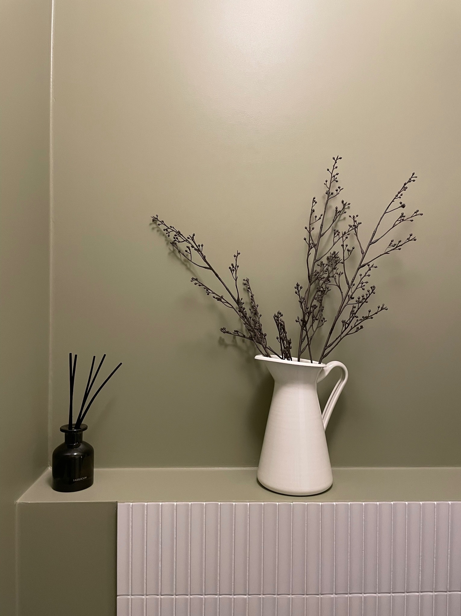 Minimalistyczna łazienka: biały dzbanek z gałązkami i czarny dyfuzor zapachowy na tle oliwkowej ściany i białych, wąskich płytek.
