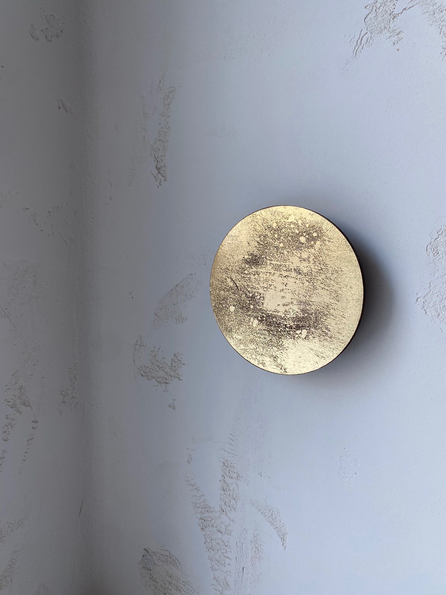 Dekoracyjna, złota rozeta na szarej ścianie z delikatną fakturą. Okrągła ozdoba z efektem przetarć, umieszczona na styku dwóch ścian, tworzy unikalny akcent wnętrza.
