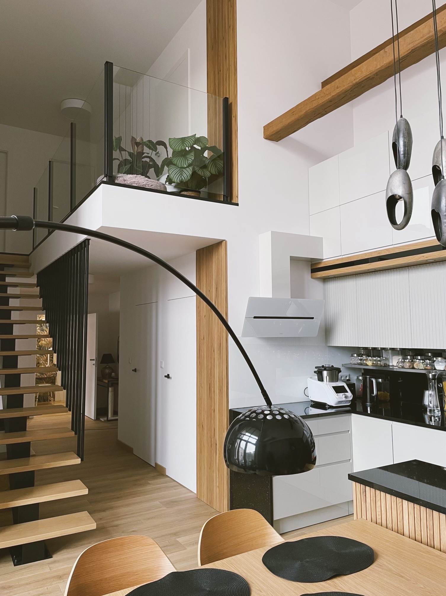 Dwupoziomowe mieszkanie: jasna kuchnia z drewnianymi akcentami, schody z czarną balustradą i antresola z roślinami. Nowoczesny wystrój.