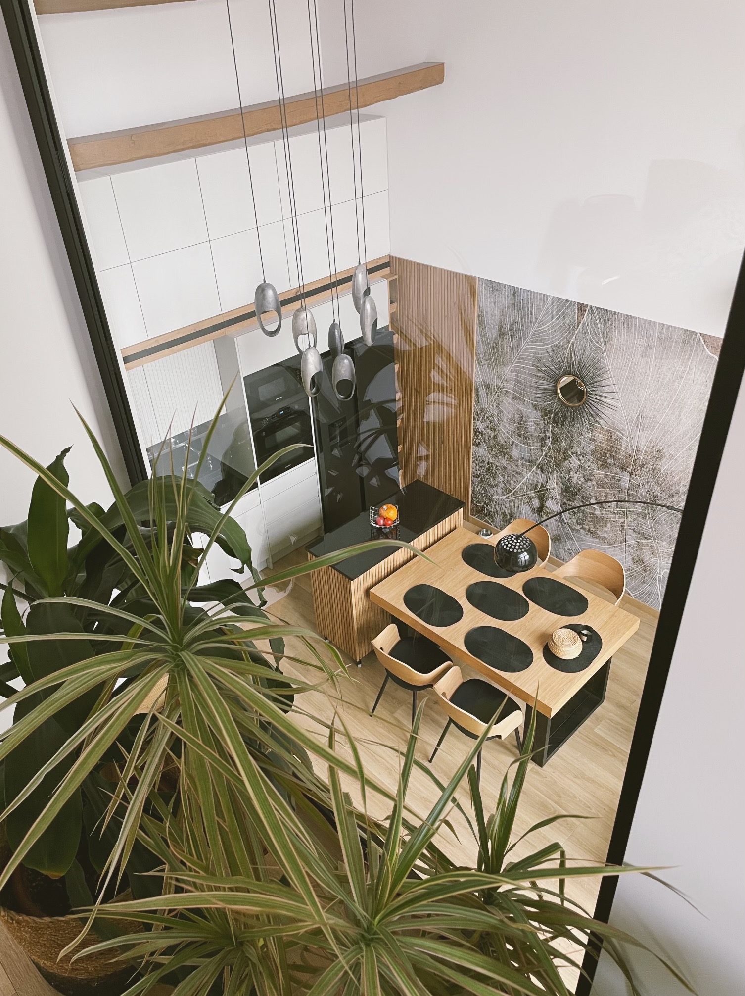Nowoczesna jadalnia z drewnianym stołem i krzesłami, widok z góry, akcent roślinny w postaci dużej rośliny doniczkowej, designerskie lampy wiszące.