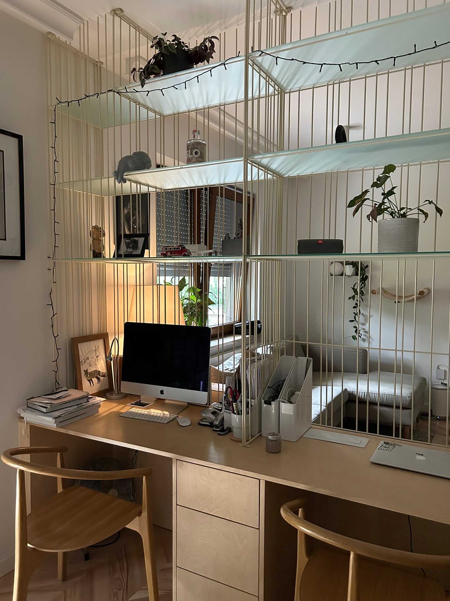 Domowe biuro z biurkiem z jasnego drewna, krzesłami i regałem z dekoracjami. Widok na pokój dzienny przez regał. Ciepłe oświetlenie.