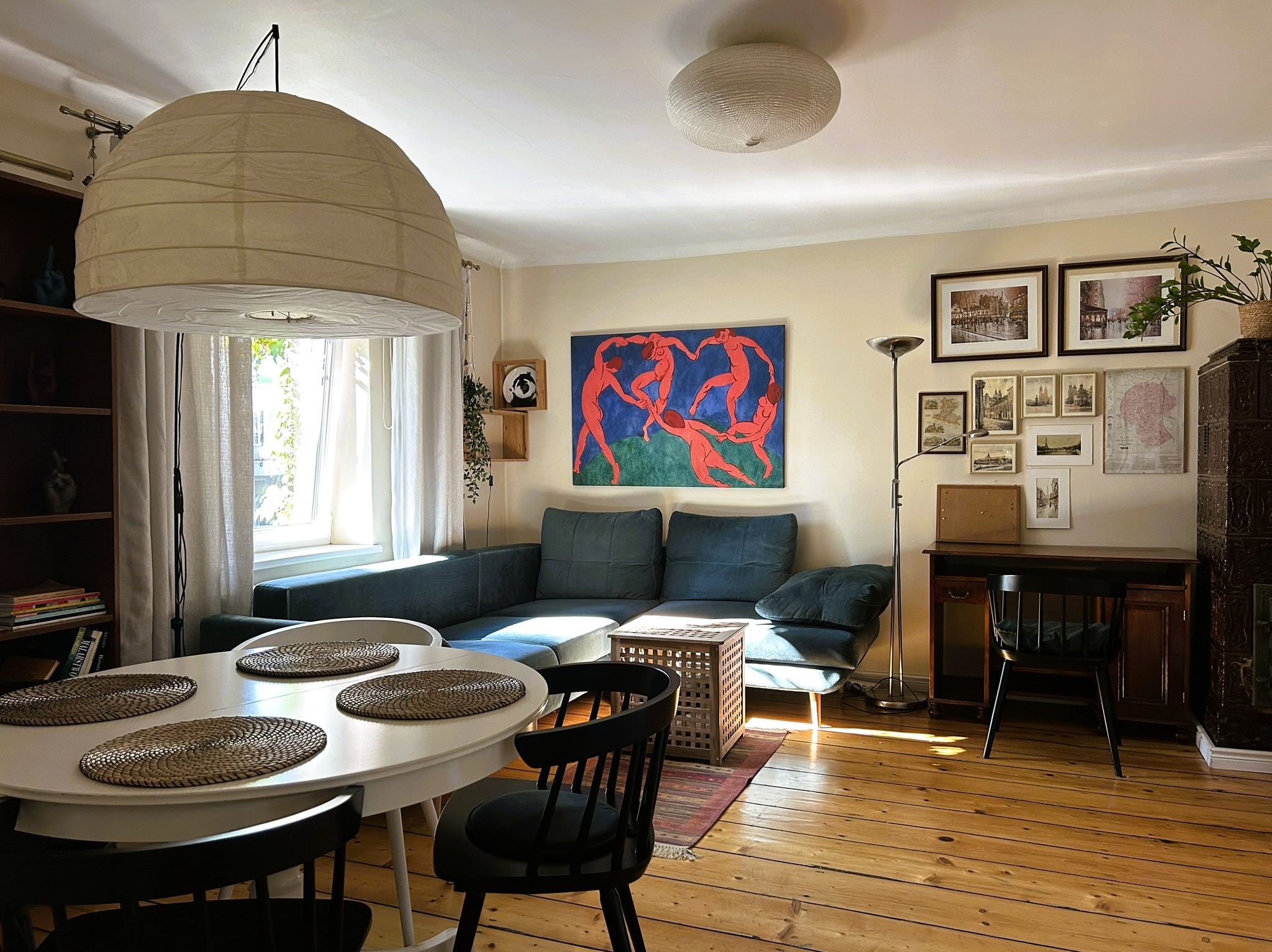 Jasne wnętrze salonu z drewnianą podłogą, niebieską sofą narożną i okrągłym stołem z czarnymi krzesłami. Na ścianie wiszą obrazy i grafiki.