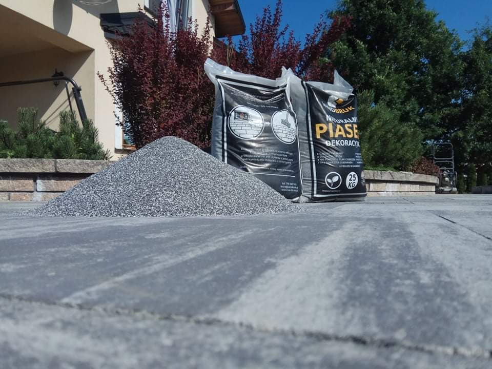 Góra szarego kruszywa na ułożonej, szarej kostce brukowej, obok dwa worki z napisem 'Piasek Dekoracyjny' i ubijarka w tle.