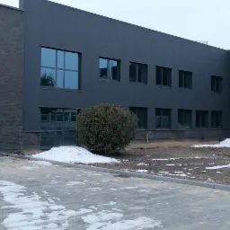 Nowoczesny budynek z szarej cegły i paneli elewacyjnych, duże okna z refleksami nieba, częściowo odśnieżony teren wokół budynku.