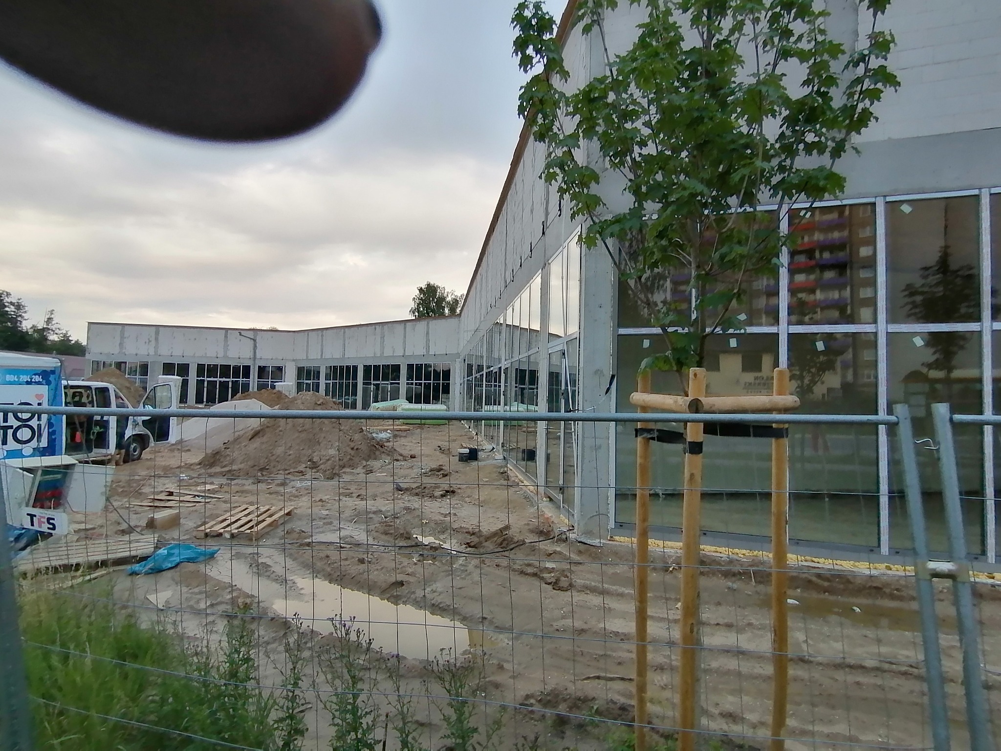 Budowa budynku z zamontowanymi oknami, widoczny teren budowy z ziemią, kałużami i ogrodzeniem tymczasowym, w tle zaparkowany samochód dostawczy.