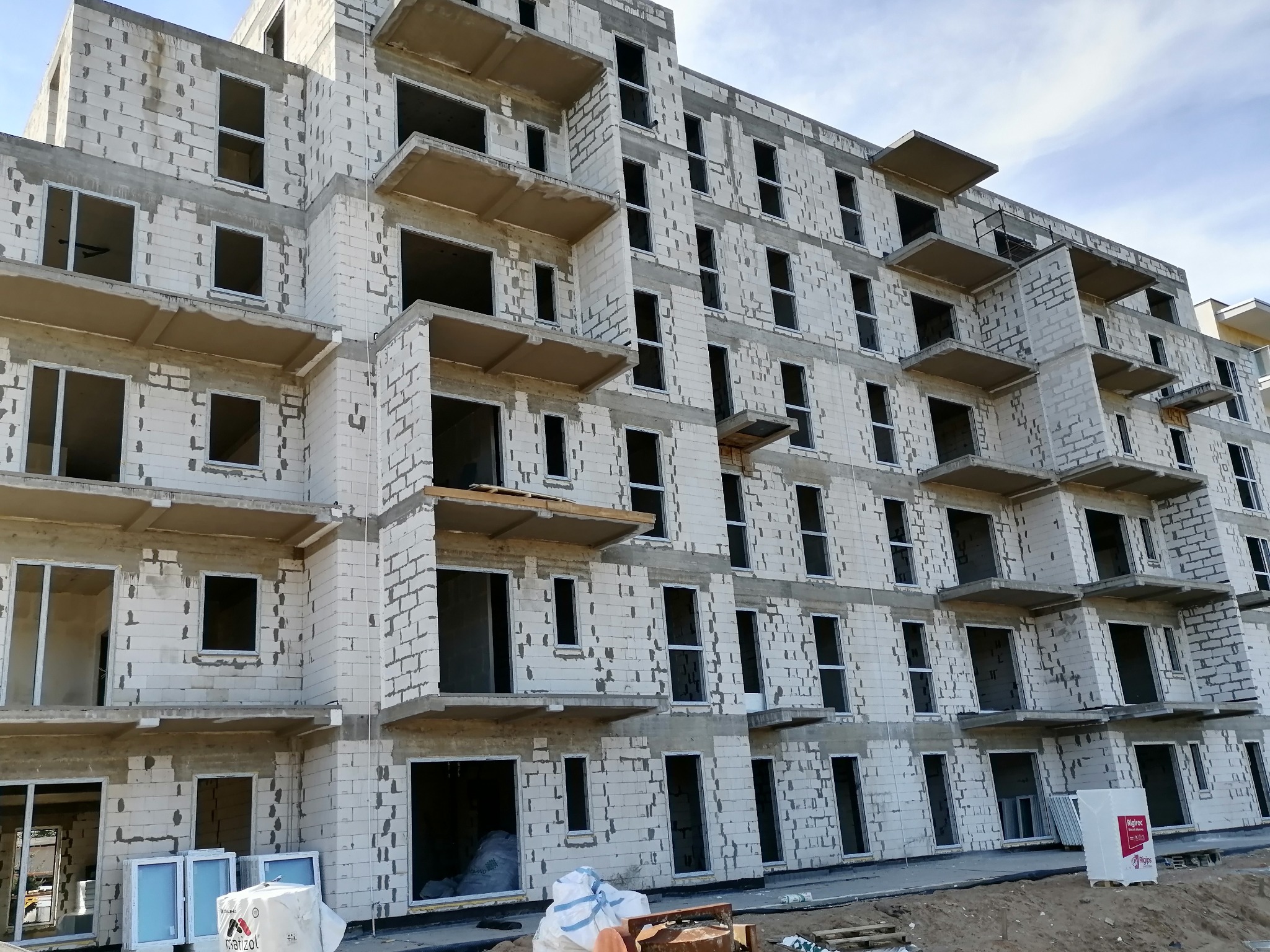 Budynek wielorodzinny w trakcie budowy, widoczne puste otwory okienne i balkonowe, przygotowane do montażu stolarki, na dole palety i materiały budowlane.