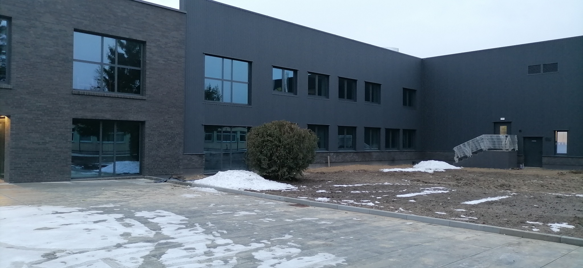 Nowoczesny budynek z szarej cegły i paneli elewacyjnych, duże okna z refleksami nieba, częściowo odśnieżony teren wokół budynku.