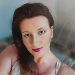 Portret kobiety z długimi, rudymi włosami, intensywnym makijażem oczu i ust w odcieniu fuksji, uśmiechająca się delikatnie.