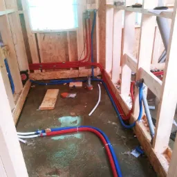 Instalacja wodno-kanalizacyjna i centralnego ogrzewania w surowym domu szkieletowym: widoczne czerwone i niebieskie rury PEX prowadzone wzdłuż drewnianych belek konstrukcyjnych, białe rury...