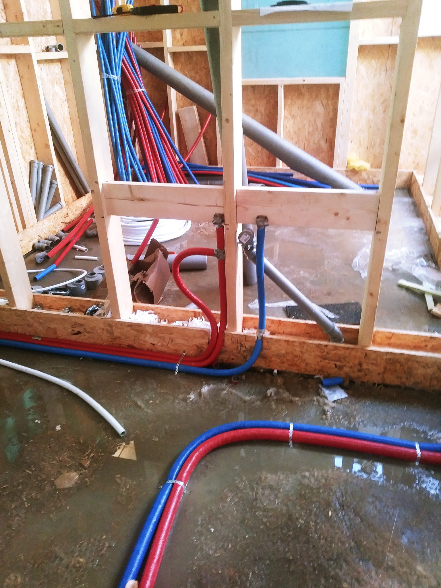 Wnętrze domu szkieletowego w trakcie prac instalacyjnych: widoczne czerwone i niebieskie rury instalacji wodnej i grzewczej przytwierdzone do drewnianej konstrukcji ściany, podłoże zalane wodą.