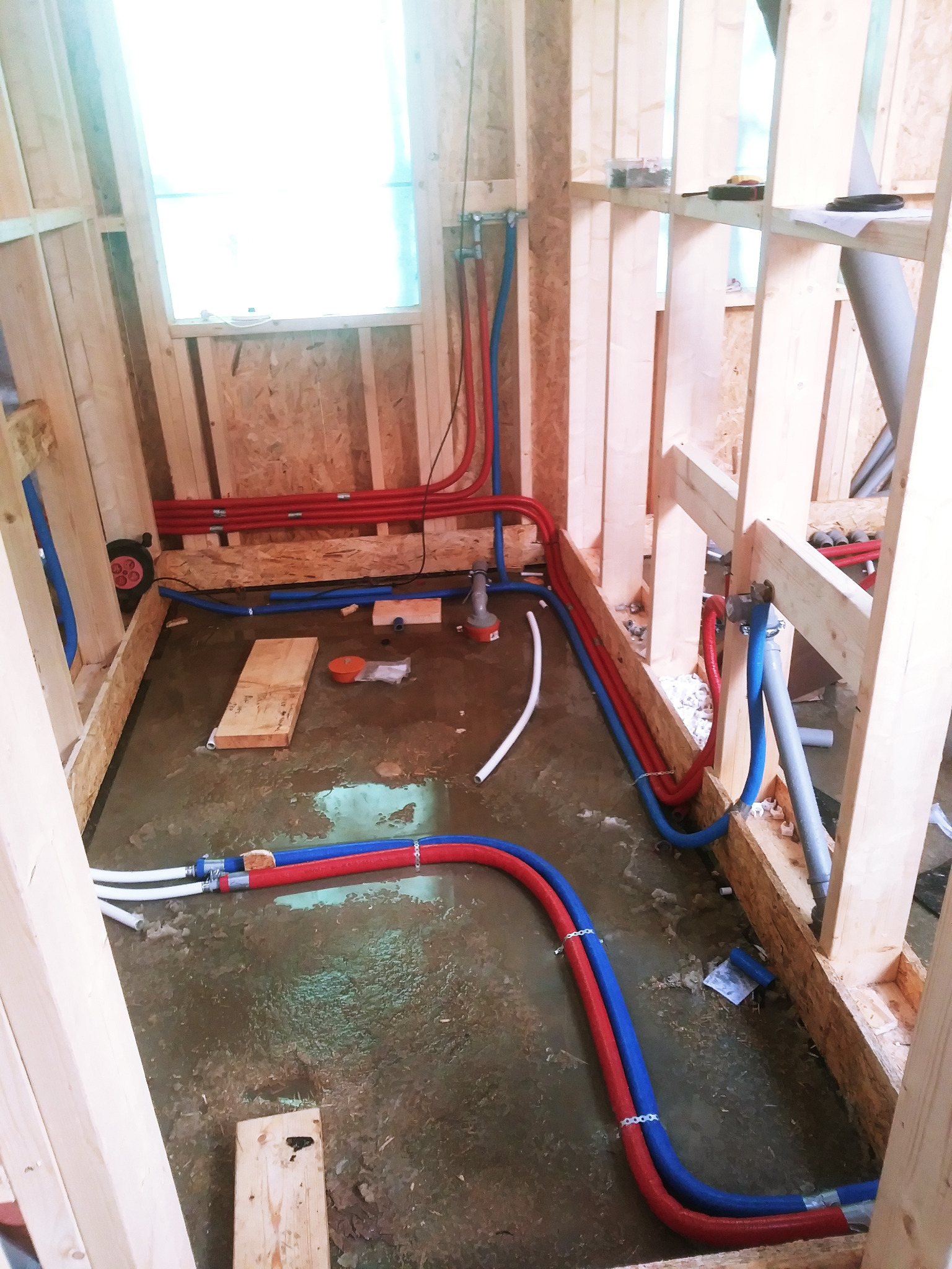 Instalacja wodno-kanalizacyjna i centralnego ogrzewania w surowym domu szkieletowym: widoczne czerwone i niebieskie rury PEX prowadzone wzdłuż drewnianych belek konstrukcyjnych, białe rury...