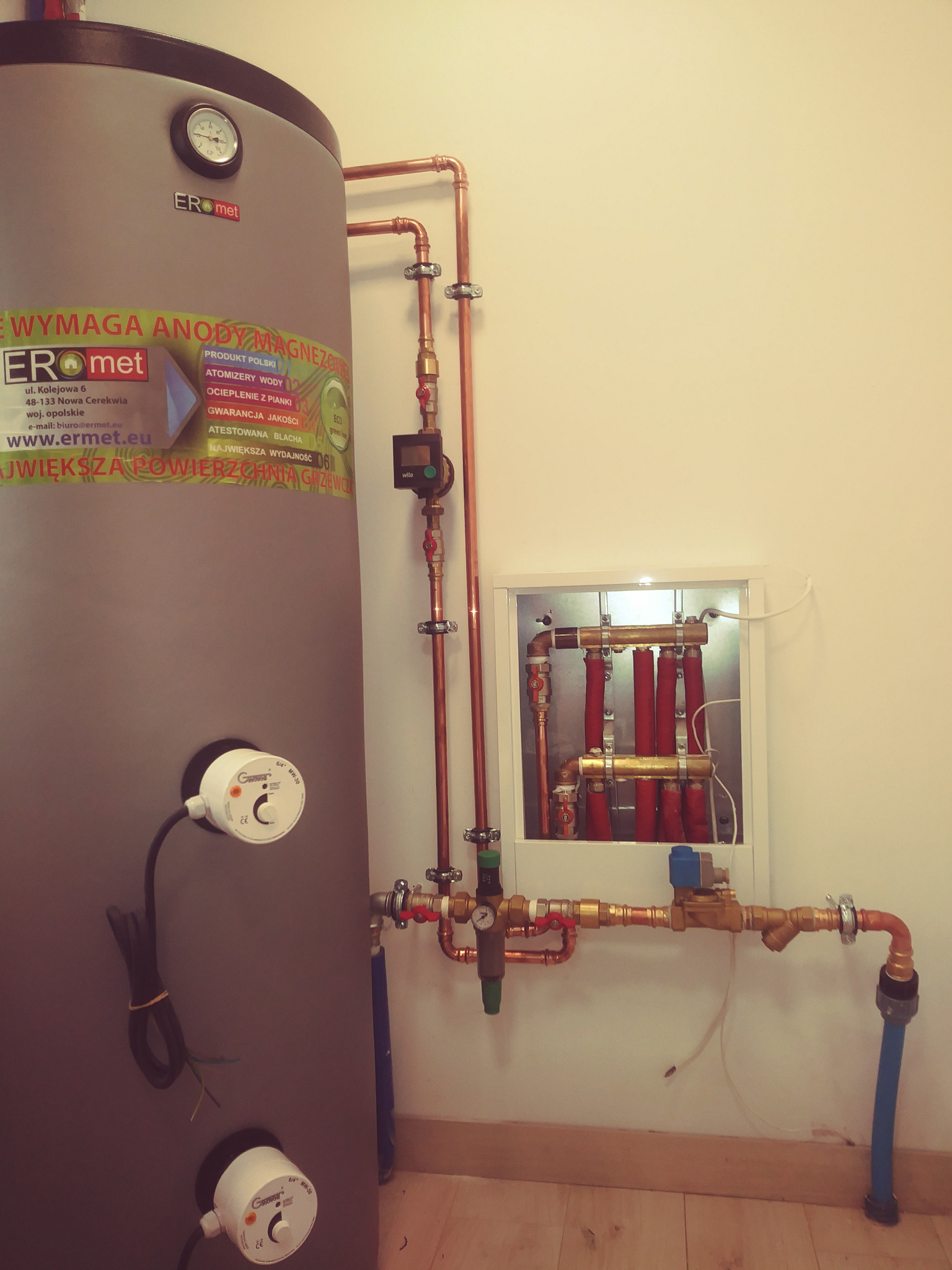 Szary bojler marki Eromet z widocznym manometrem i naklejką informacyjną, połączony miedzianymi rurami z rozdzielaczem w białej obudowie oraz z zaworami i niebieską rurą doprowadzającą wodę.