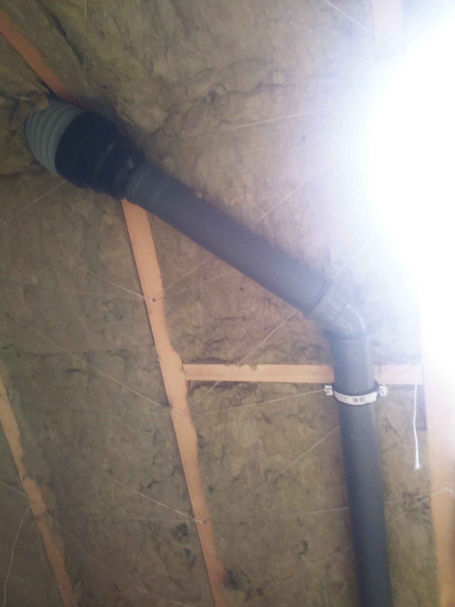 Czarna rura kanalizacyjna z elastycznym łącznikiem przymocowana do drewnianej konstrukcji dachu ocieplonego wełną mineralną, zabezpieczoną sznurkiem.