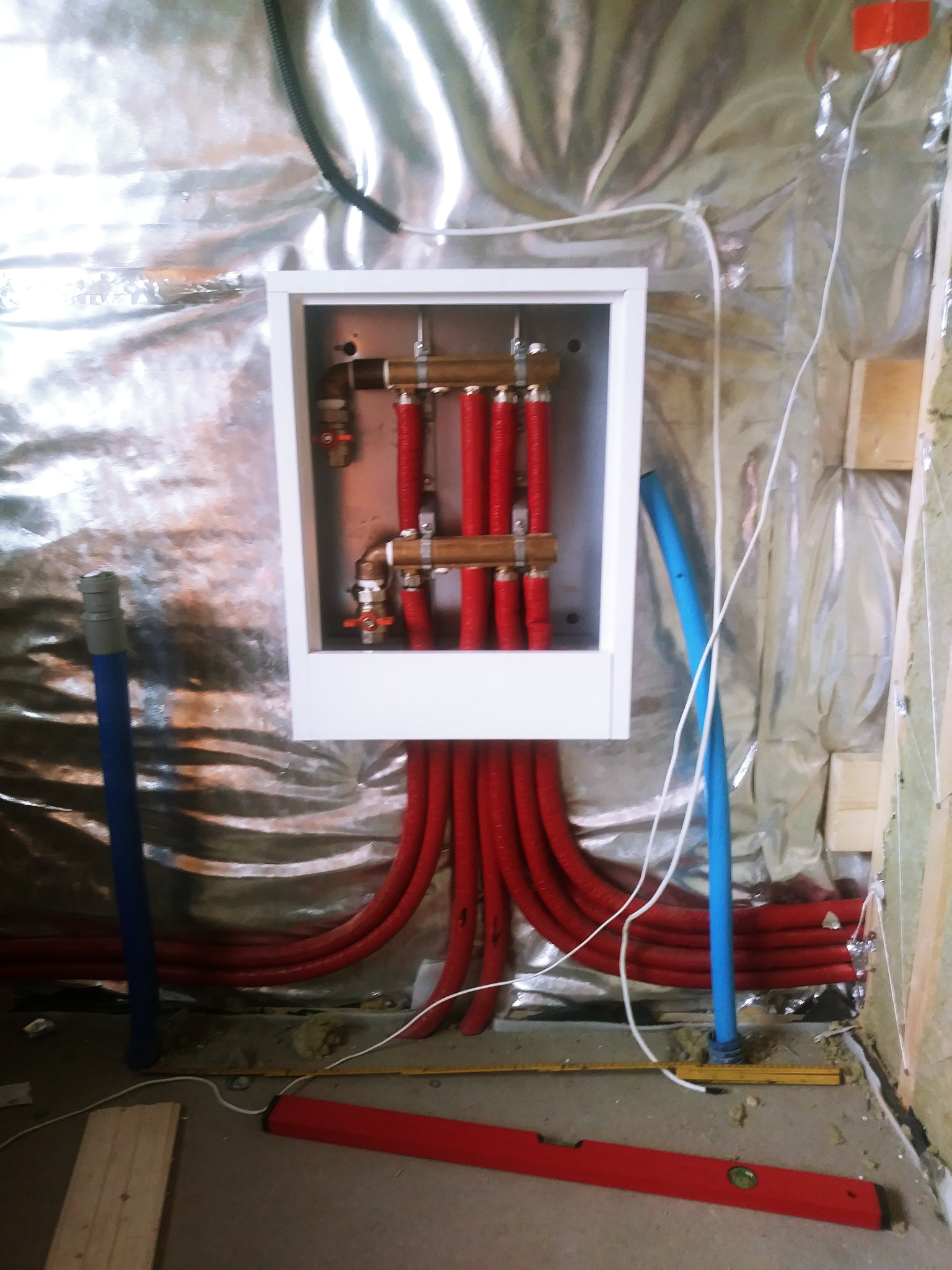 Rozdzielacz centralnego ogrzewania w białej obudowie, z czerwonymi rurami PEX, zamontowany na ścianie z izolacją z folii aluminiowej w domu szkieletowym, widoczny poziomica i fragment niebieskiej...