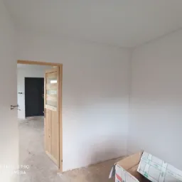 Świeżo pomalowane na biało wnętrze pokoju z widocznymi dwoma ościeżnicami drzwiowymi, karton na podłodze i surowa podłoga.