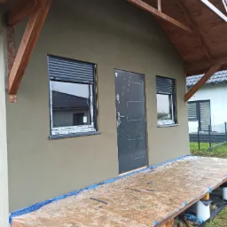 Elewacja budynku po nałożeniu szarej warstwy, widoczne okna z roletami zewnętrznymi, stalowe drzwi wejściowe, drewniana konstrukcja dachu i prowizoryczny podest.