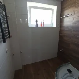 Wykończona łazienka z białymi i drewnopodobnymi płytkami, grzejnikiem drabinkowym po lewej stronie, oknem z kosmetykami i częścią kabiny prysznicowej w prawym dolnym rogu.