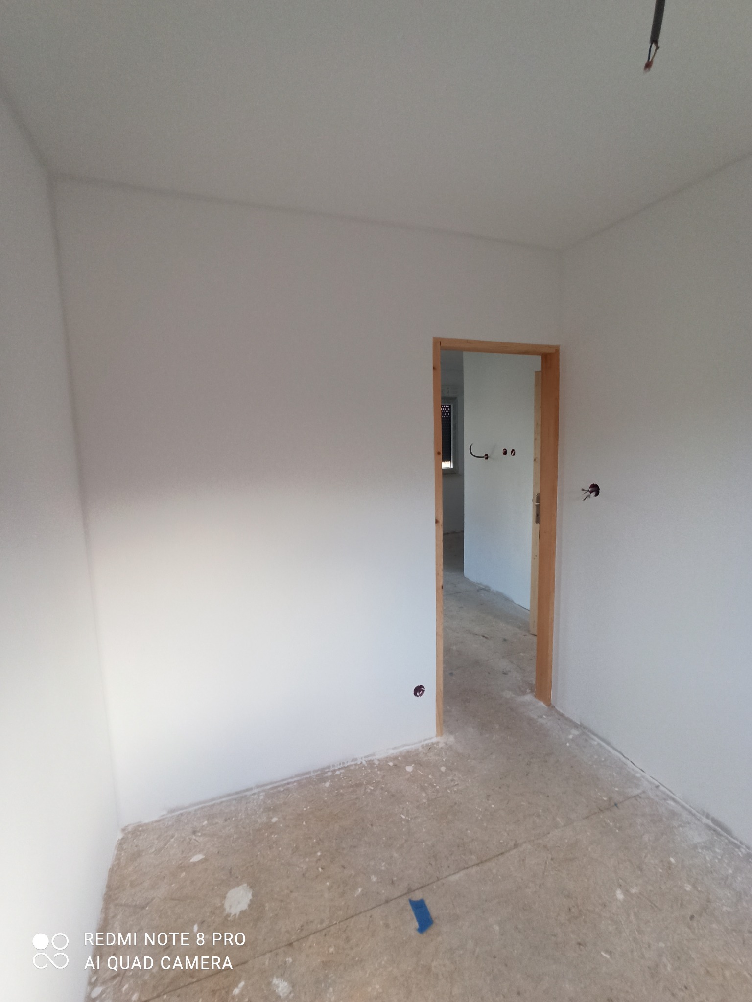 Wnętrze pokoju po malowaniu ścian na biało, widoczny fragment podłogi zabezpieczonej folią malarską i otwór drzwiowy z drewnianą ościeżnicą.