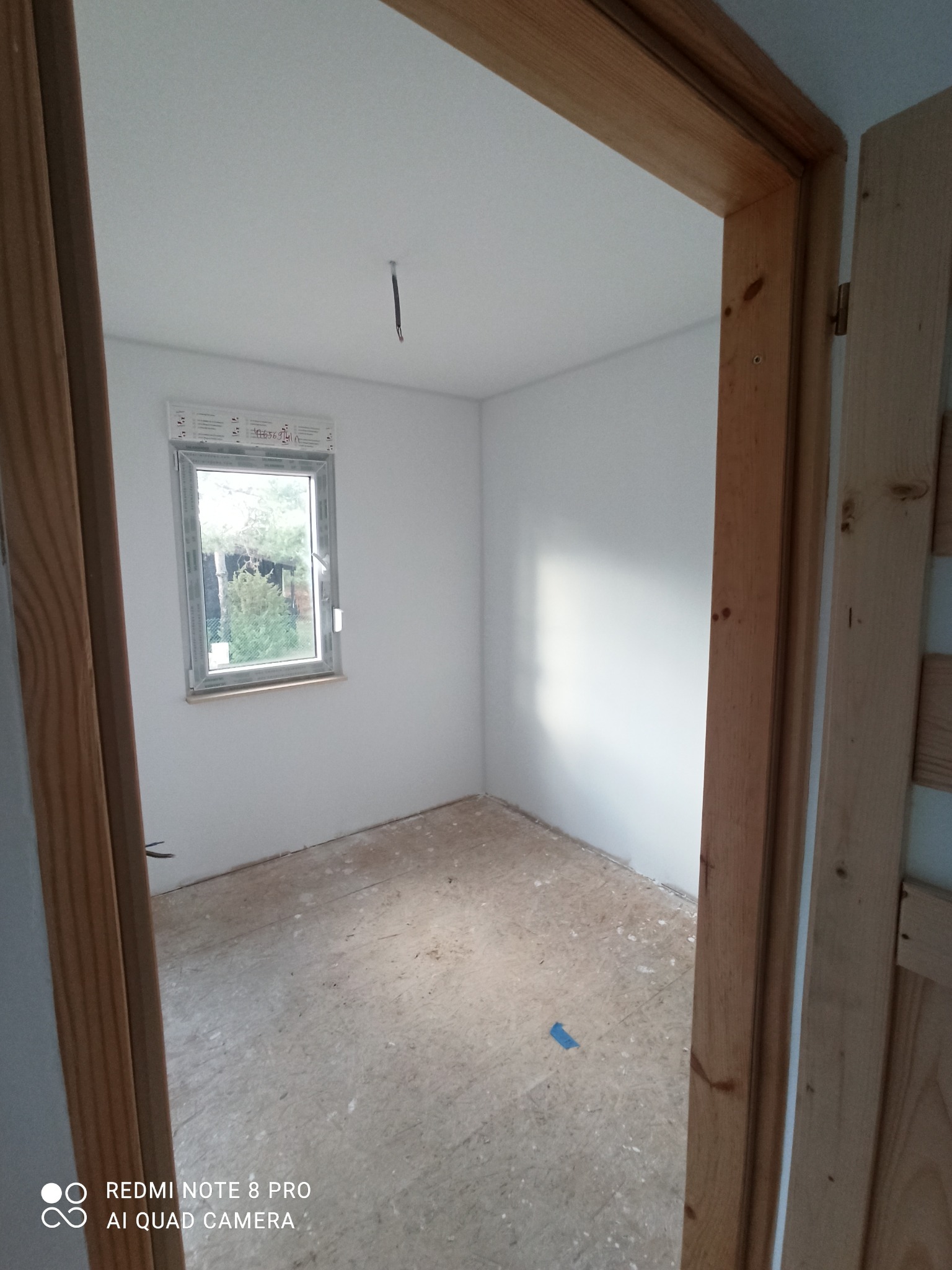 Pokój po świeżym odmalowaniu na biało, widoczne surowe drewniane ościeżnice drzwi i okna, na podłodze widoczne pozostałości po pracach remontowych.