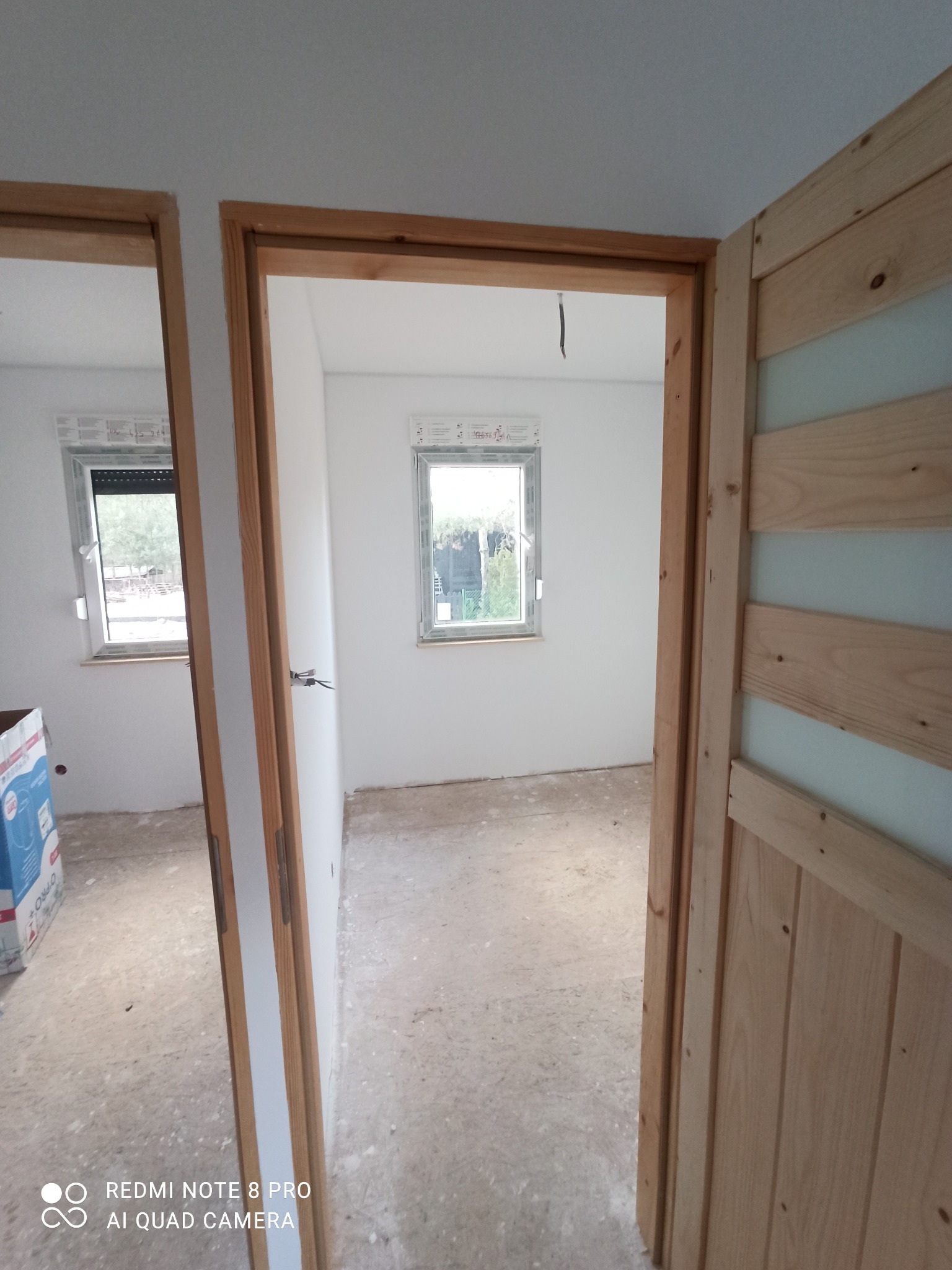 Wnętrze pokoju w trakcie prac wykończeniowych, widoczne dwa okna i ościeżnice drzwiowe z surowego drewna, podłoga przygotowana do dalszych prac, ściany po wstępnym przygotowaniu.