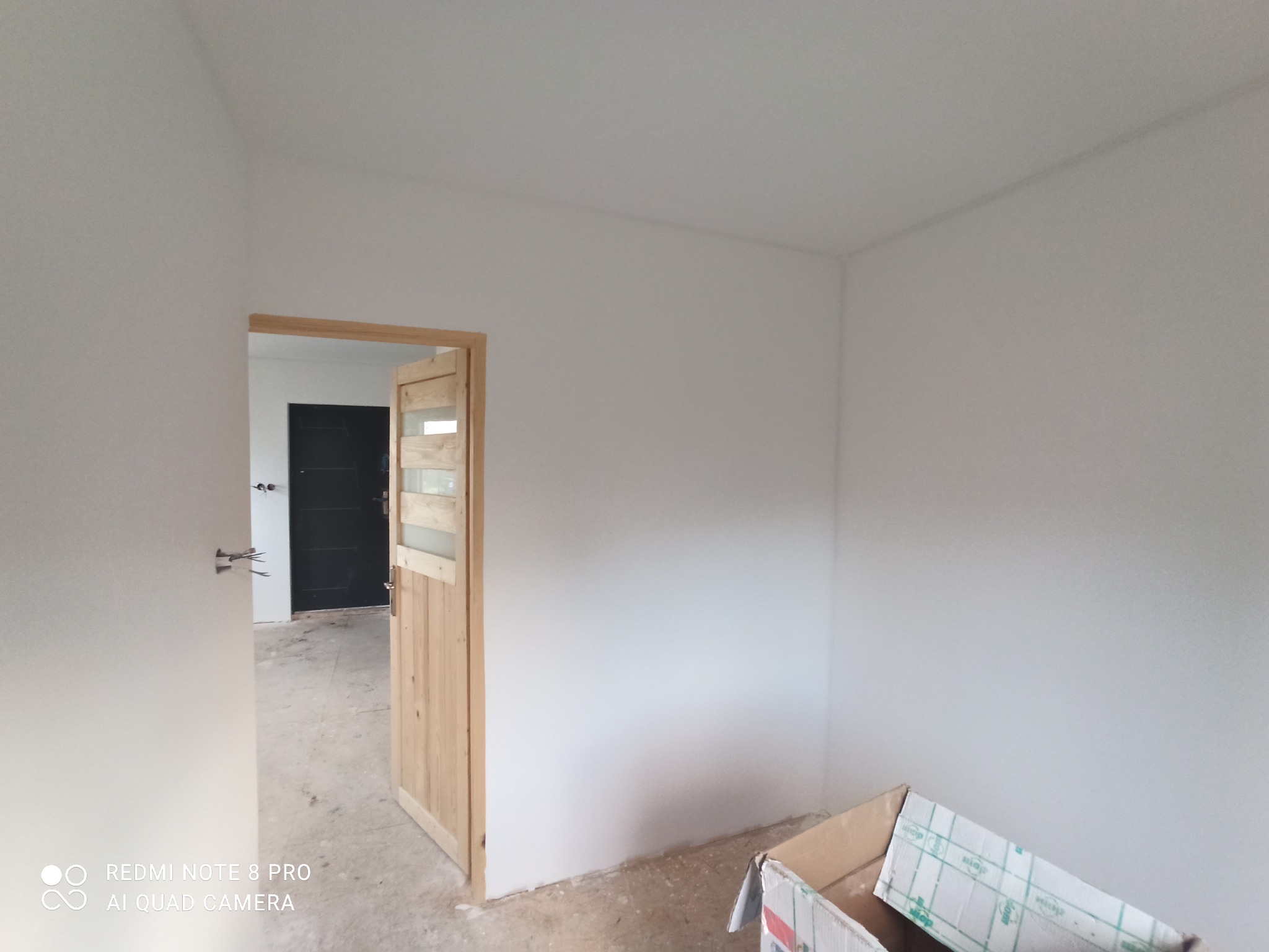 Świeżo pomalowane na biało wnętrze pokoju z widocznymi dwoma ościeżnicami drzwiowymi, karton na podłodze i surowa podłoga.