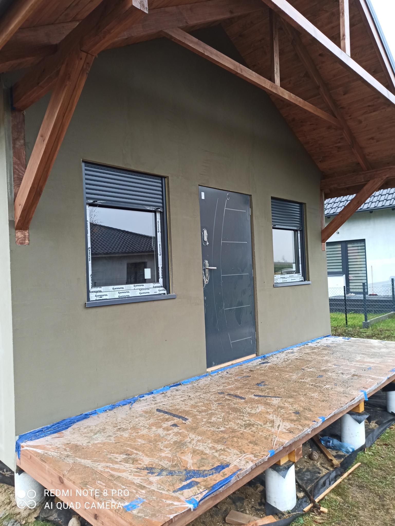 Elewacja budynku po nałożeniu szarej warstwy, widoczne okna z roletami zewnętrznymi, stalowe drzwi wejściowe, drewniana konstrukcja dachu i prowizoryczny podest.