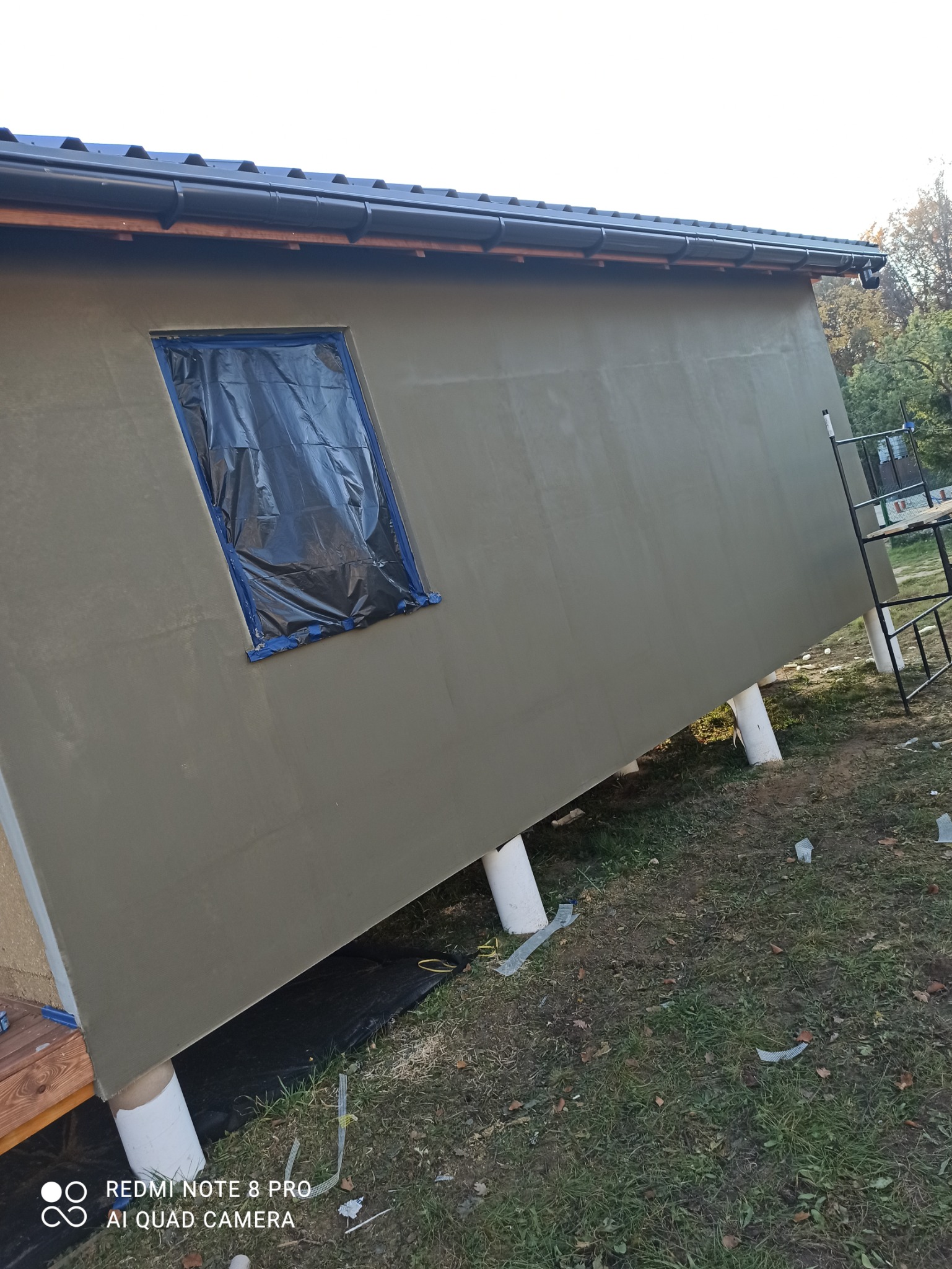 Ściana budynku po nałożeniu warstwy szarej zaprawy, okno zabezpieczone folią, widoczne rury podtrzymujące konstrukcję i rusztowanie z boku