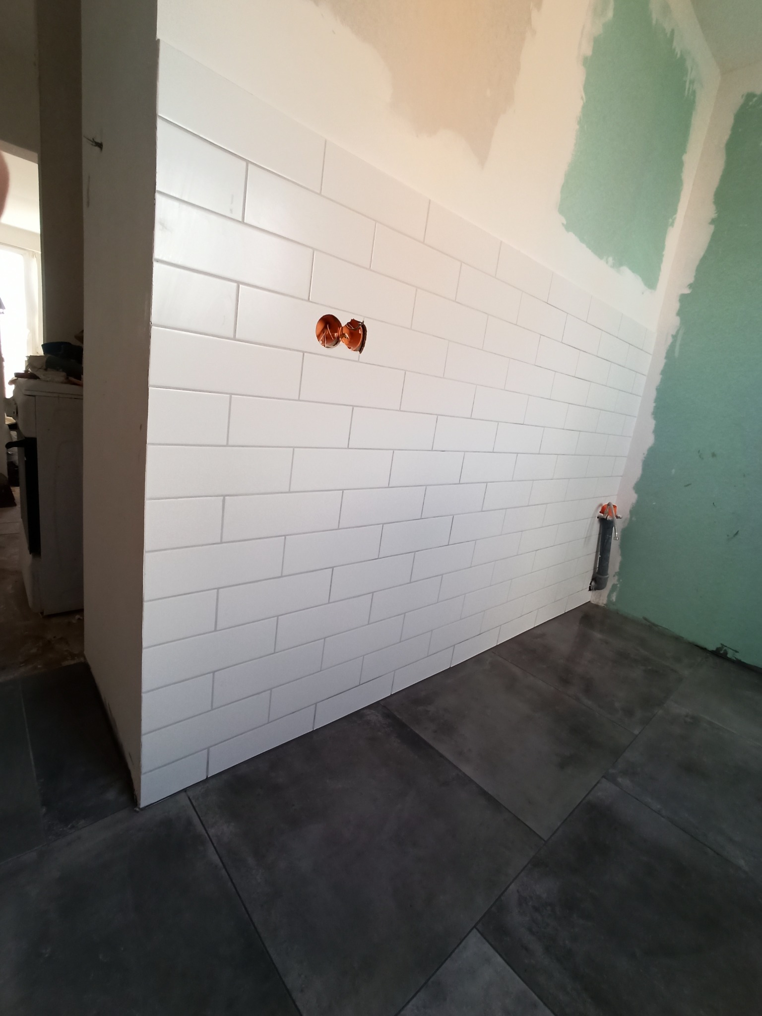 Białe, prostokątne płytki ułożone na ścianie, widoczne gniazdka elektryczne i fragment surowej ściany z zielonymi plamami oraz ciemna podłoga z dużych płytek.