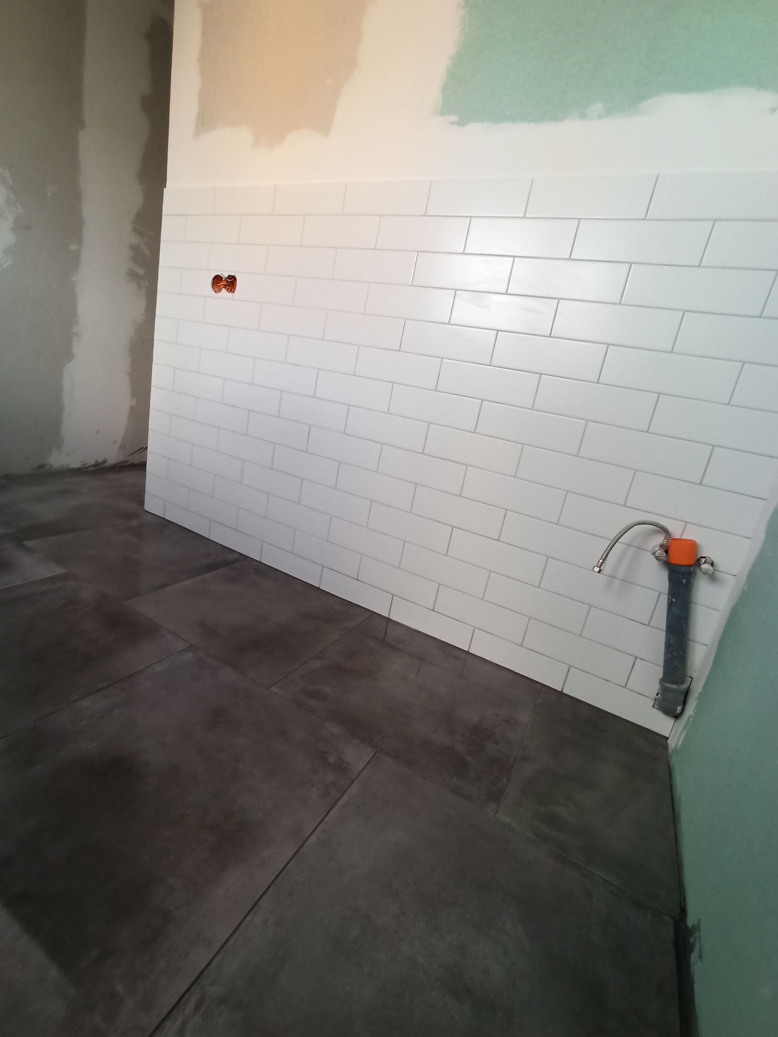 Ściana wyłożona białymi, prostokątnymi płytkami w stylu metra, ciemnoszara podłoga z płytek, widoczne przyłącze wodne z pomarańczowym elementem, fragment ściany przygotowywanej do wykończenia.