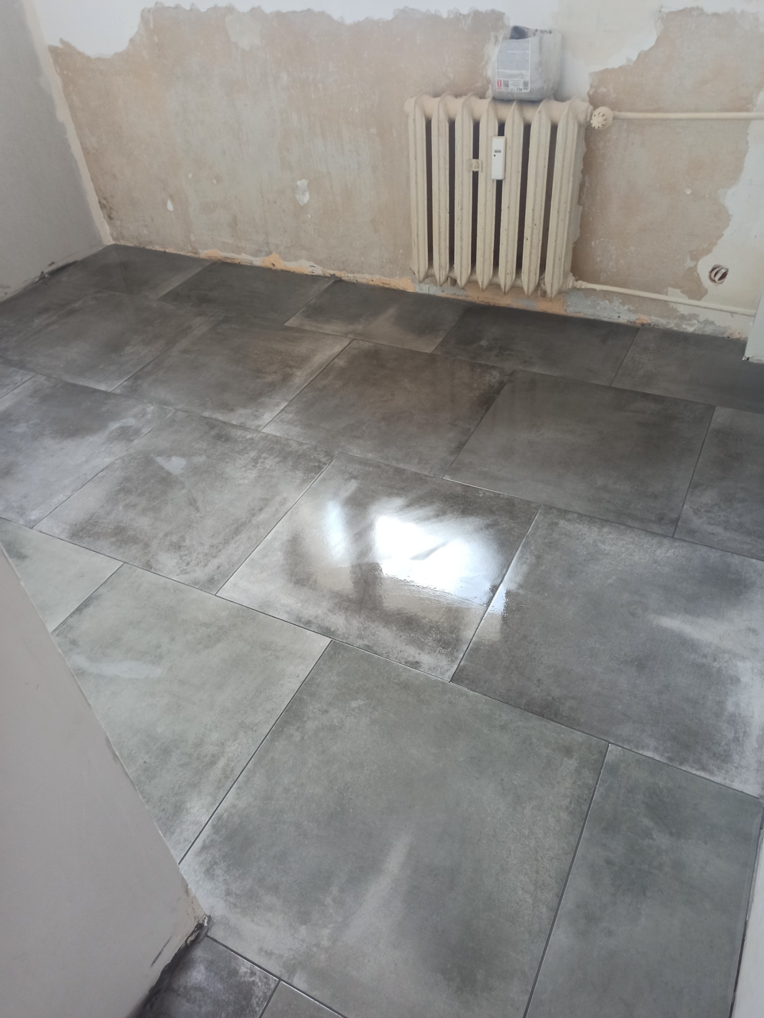 Ułożone szare płytki podłogowe w pomieszczeniu w trakcie remontu w Czarnem, widoczny grzejnik i ślady po usuniętych starych powłokach na ścianach.