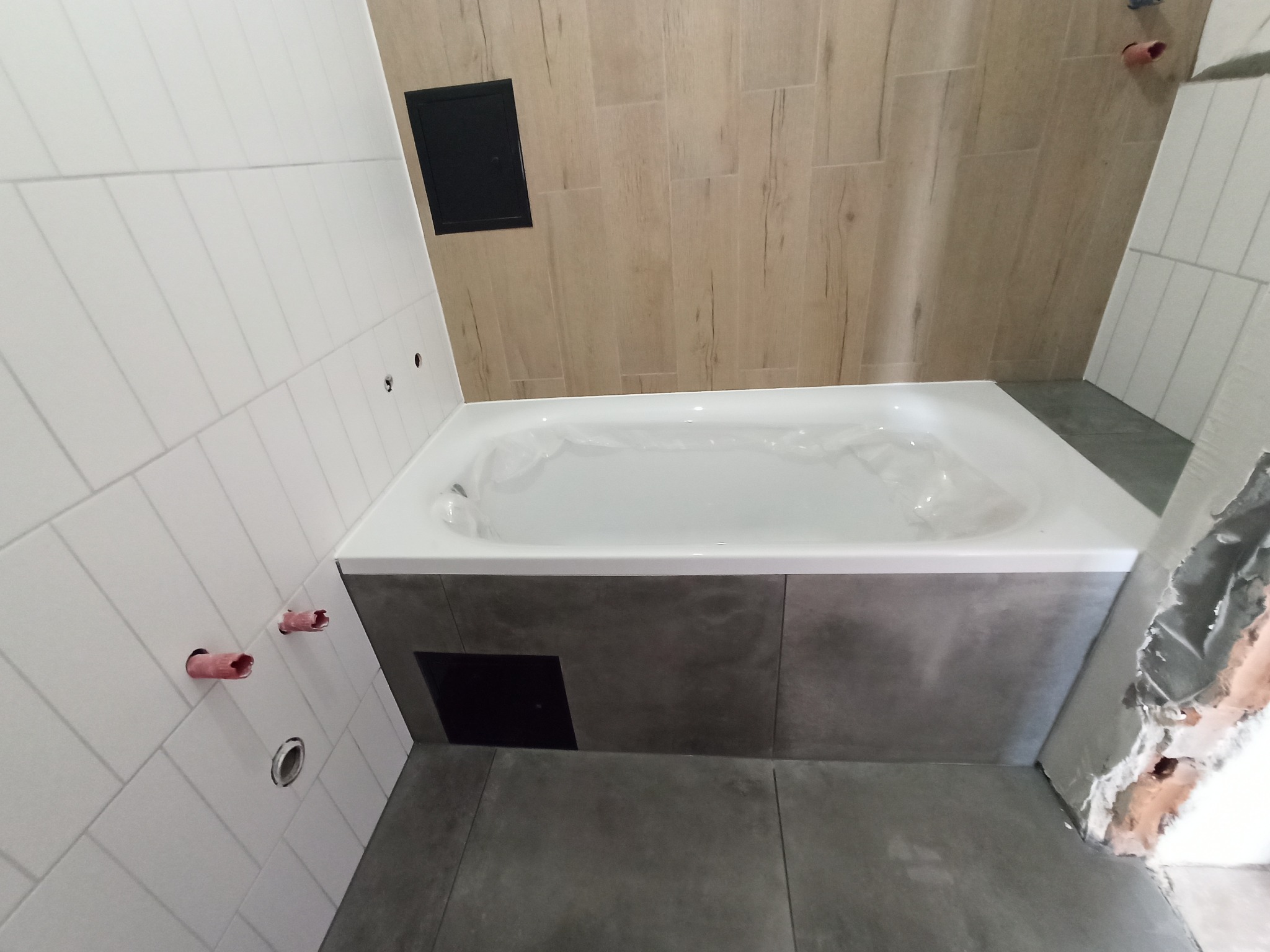 Wanna obudowana szarą mozaiką w trakcie montażu, widoczne fragmenty niezabezpieczonych rur i otwory w białych płytkach ściennych, fragment ściany w płytkach imitujących drewno.