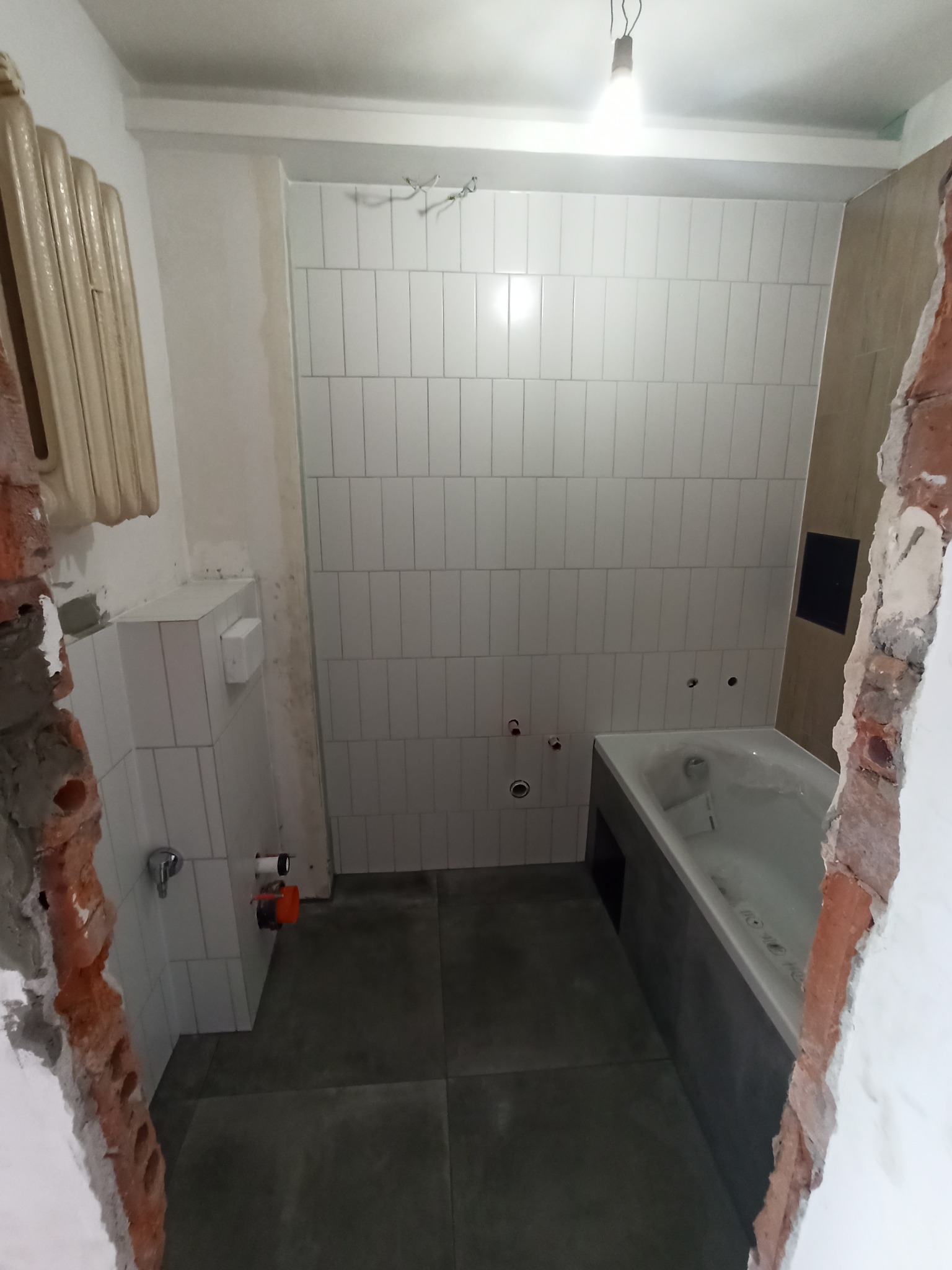 Wnętrze łazienki w trakcie prac: białe, pionowo ułożone płytki na ścianach, szara podłoga, wanna z szarą obudową, widoczne fragmenty odsłoniętej cegły przy futrynie, grzejnik żeberkowy na ścianie.