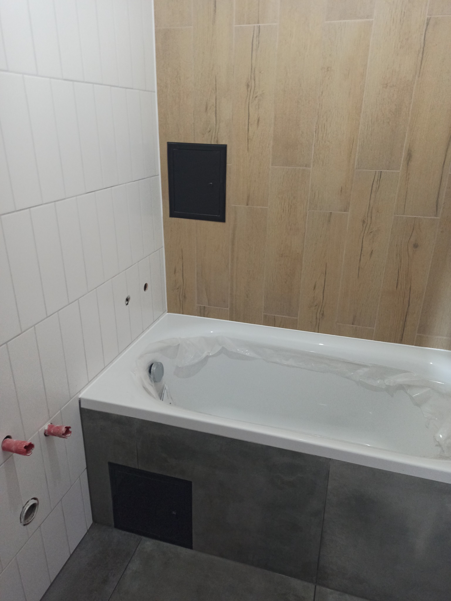 Nowa, biała wanna, zabezpieczona folią ochronną, w trakcie montażu w łazience z częściowo wykończonymi ścianami z płytek imitujących drewno i białych płytek.