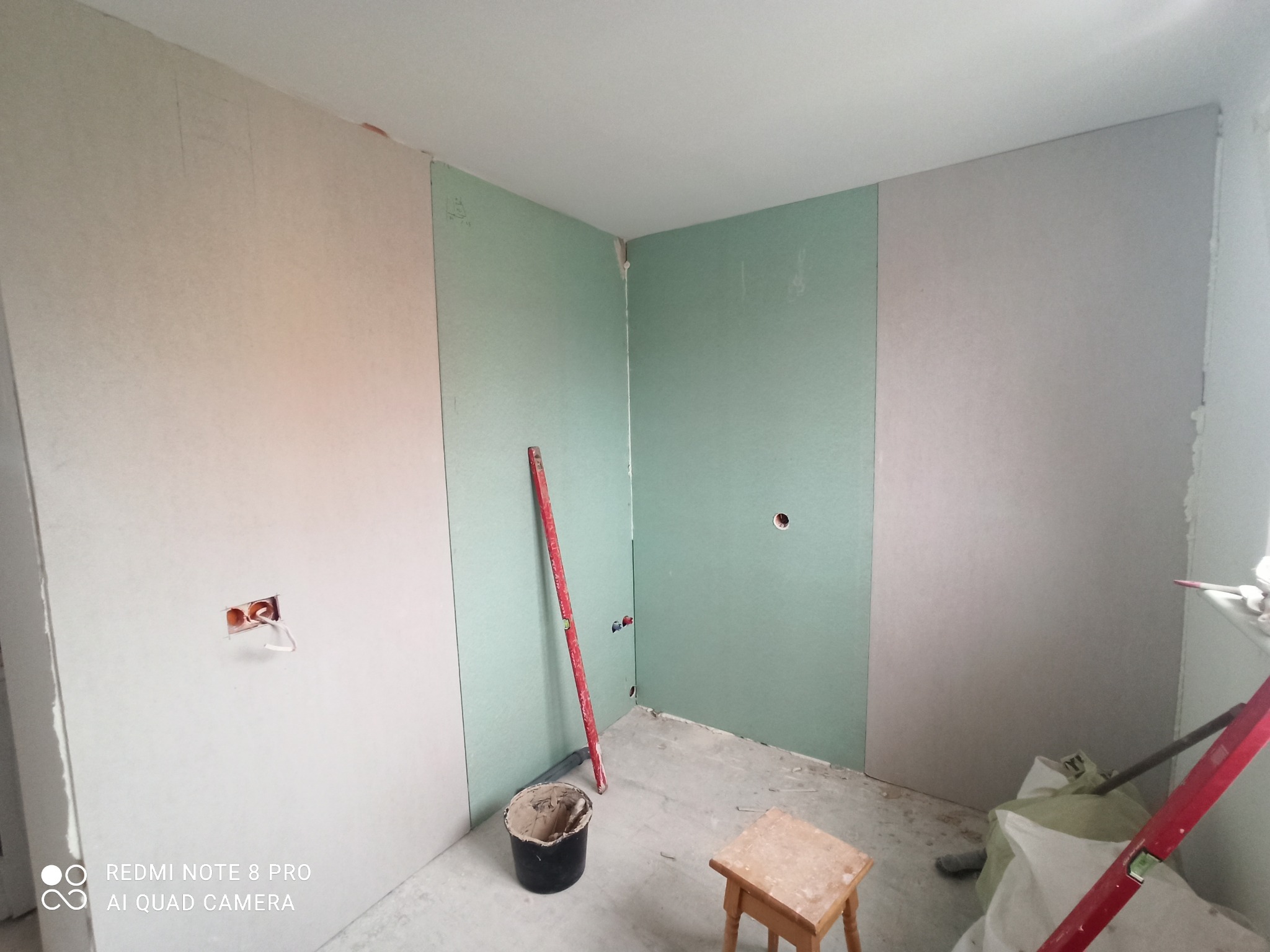 Montaż płyt gipsowo-kartonowych na ścianach pomieszczenia w trakcie prac wykończeniowych, widoczny poziomica, wiadro z zaprawą, oraz otwór na gniazdko elektryczne z wystającymi kablami.