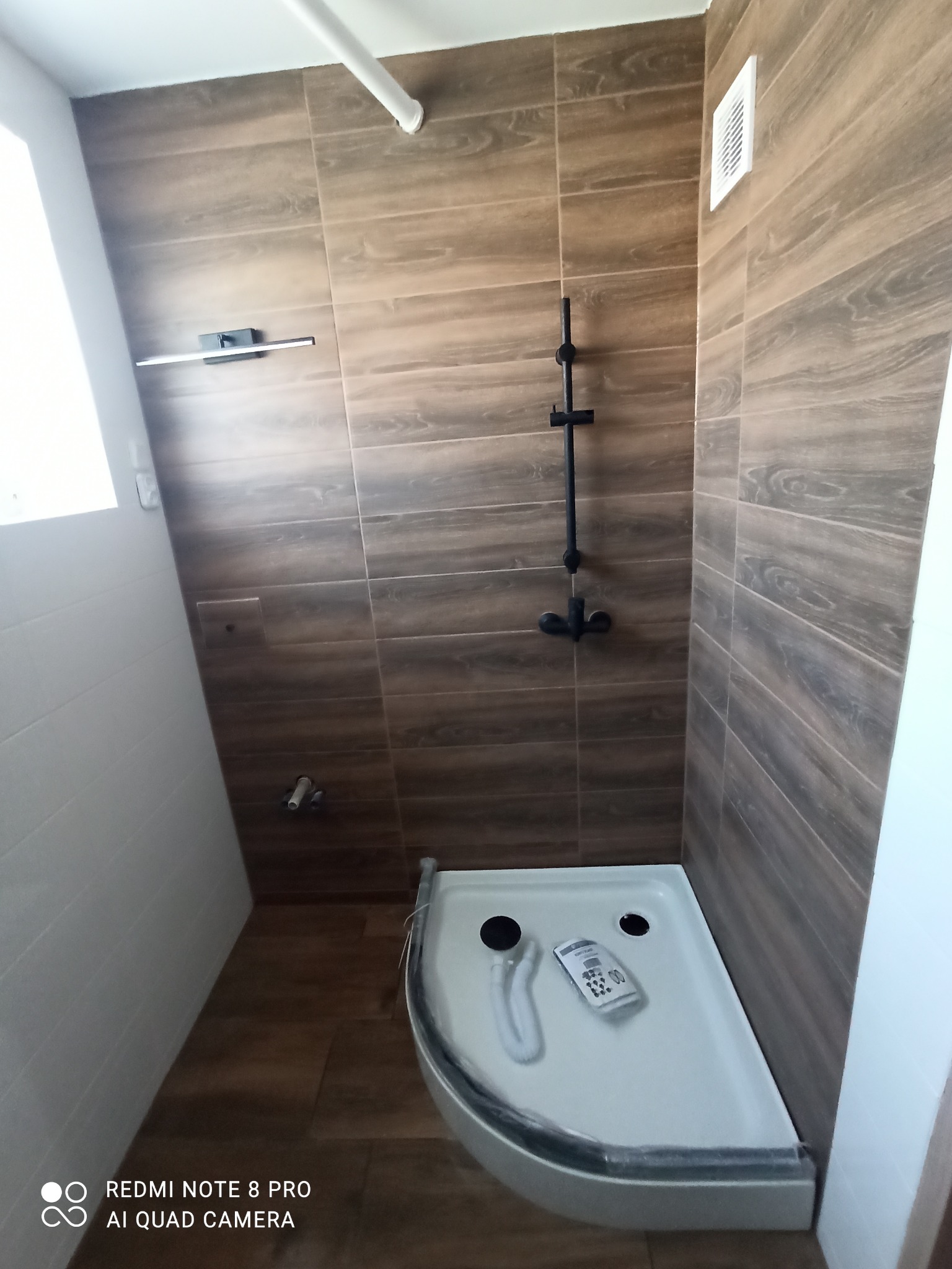 Wykończona kabina prysznicowa z brązowymi płytkami imitującymi drewno, czarna armatura, biała wentylacja i widoczne akcesoria montażowe w brodziku.