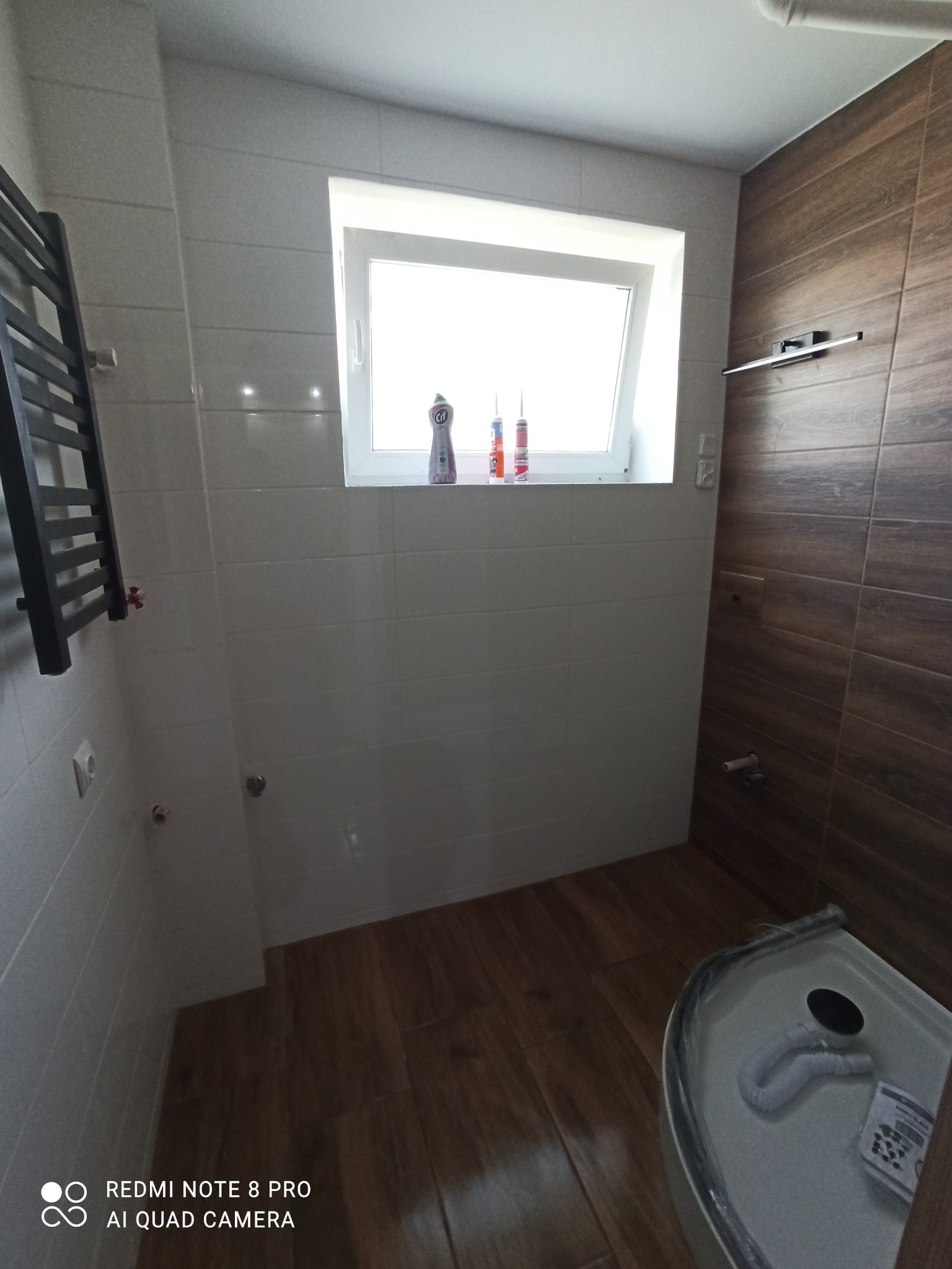 Wykończona łazienka z białymi i drewnopodobnymi płytkami, grzejnikiem drabinkowym po lewej stronie, oknem z kosmetykami i częścią kabiny prysznicowej w prawym dolnym rogu.