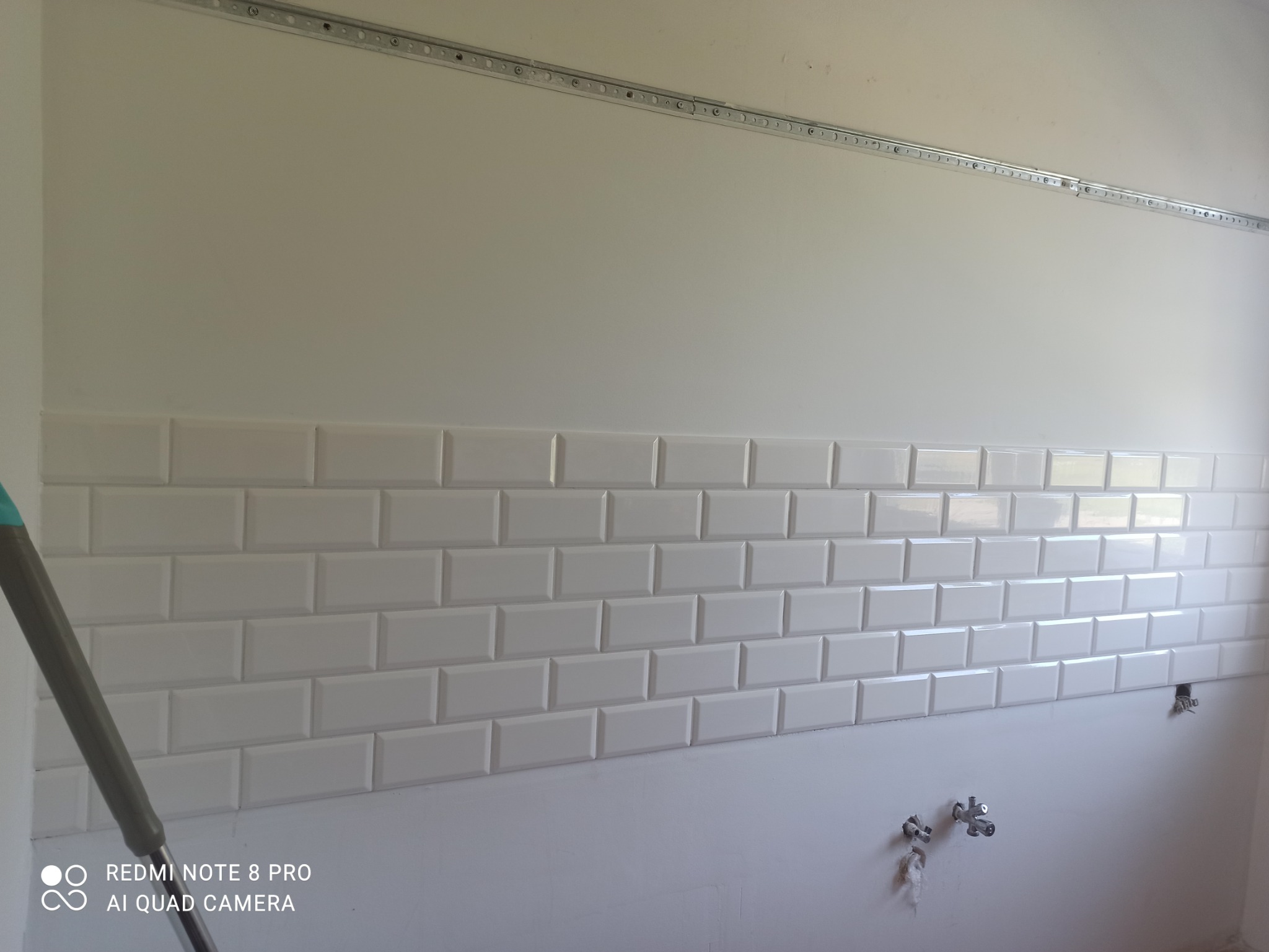 Ściana w trakcie układania białych, prostokątnych płytek typu metro, widoczne wyprowadzenia instalacji wodnej i listwa montażowa pod sufitem.