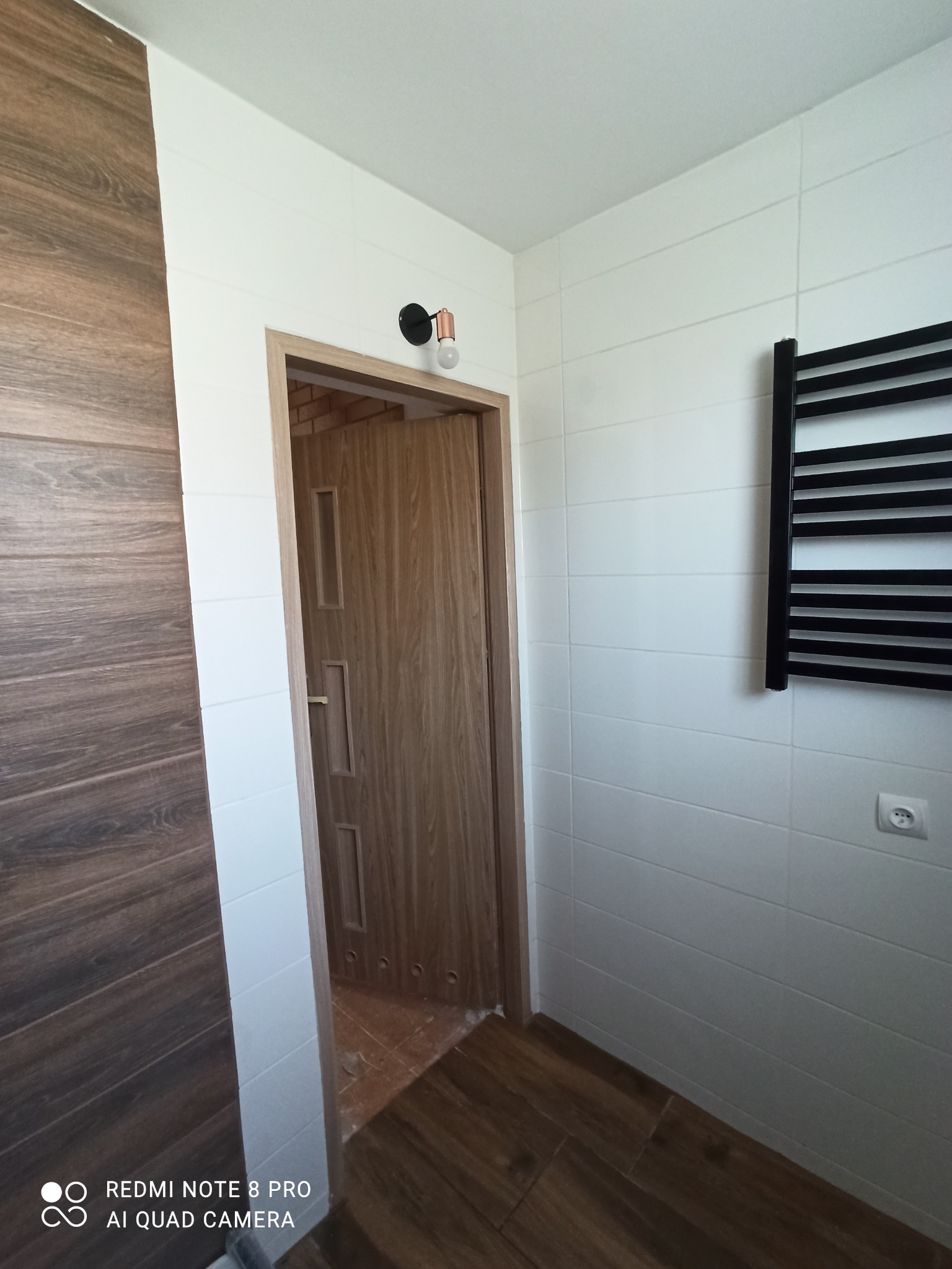 Wykończona łazienka z płytkami imitującymi drewno na ścianie i podłodze, białe płytki na ścianach, czarny grzejnik drabinkowy i drewniane drzwi z ościeżnicą.