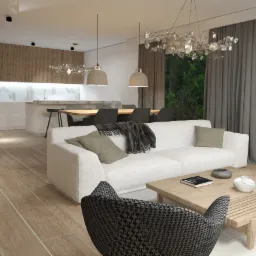 Jasne wnętrze salonu z otwartą kuchnią, biała sofa z poduszkami i pledem, drewniany stolik kawowy z książką i dekoracjami, wzorzysty fotel na pierwszym planie, lampy wiszące nad stołem jadalnym...