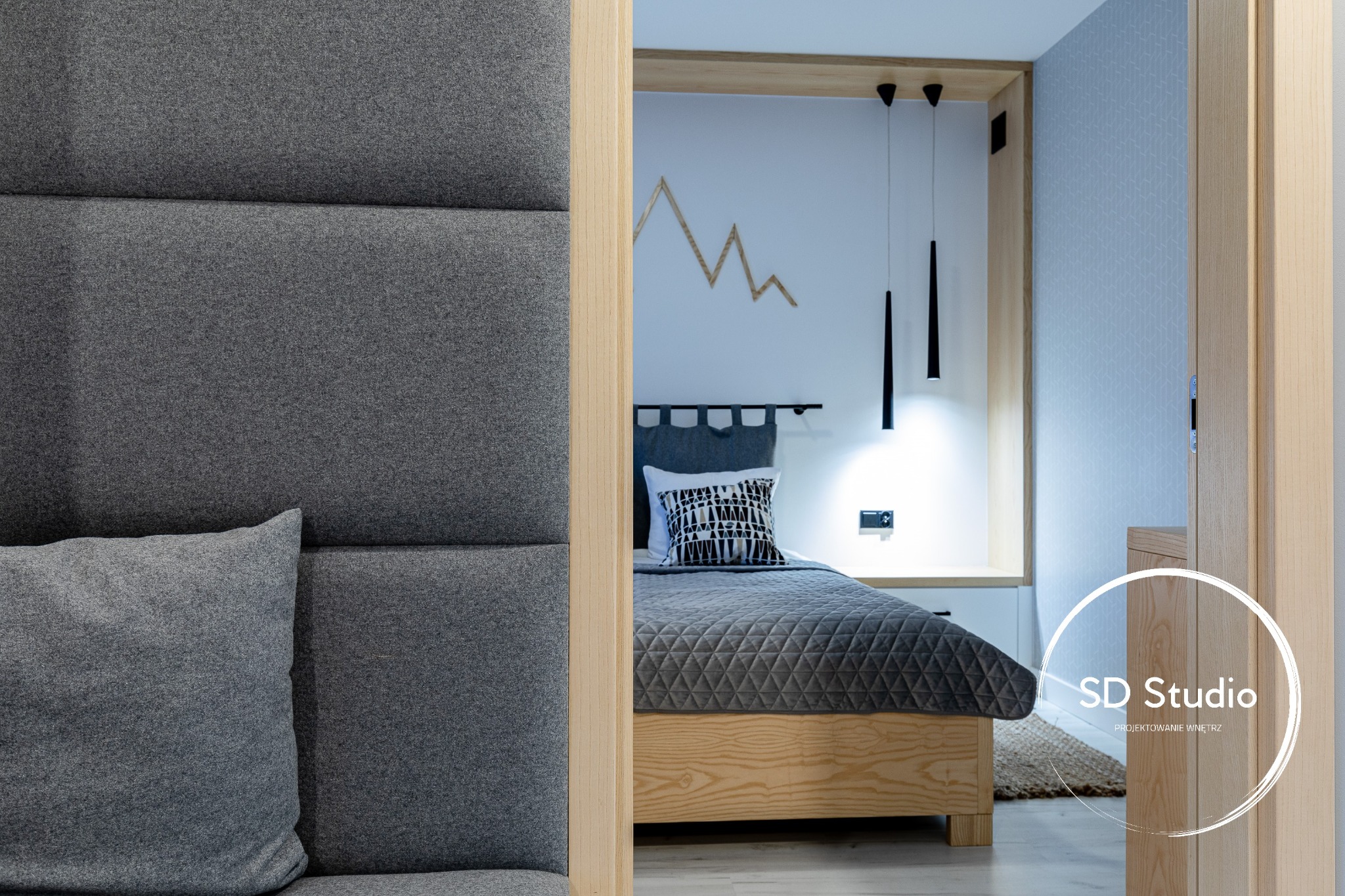 Fragment sypialni z wezgłowiem obitym szarą tkaniną, w tle łóżko z szarą narzutą, drewniana dekoracja ścienna w kształcie gór i wiszące czarne lampy, widok zza drewnianej framugi drzwi, w rogu logo...