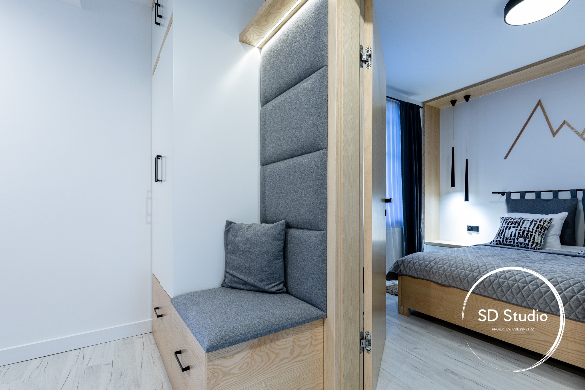 Wnęka z siedziskiem i tapicerowanym oparciem w odcieniach szarości, z drewnianą obudową i szufladami, widok na sypialnię z łóżkiem, drewnianymi elementami i dekoracją ścienną w kształcie gór.