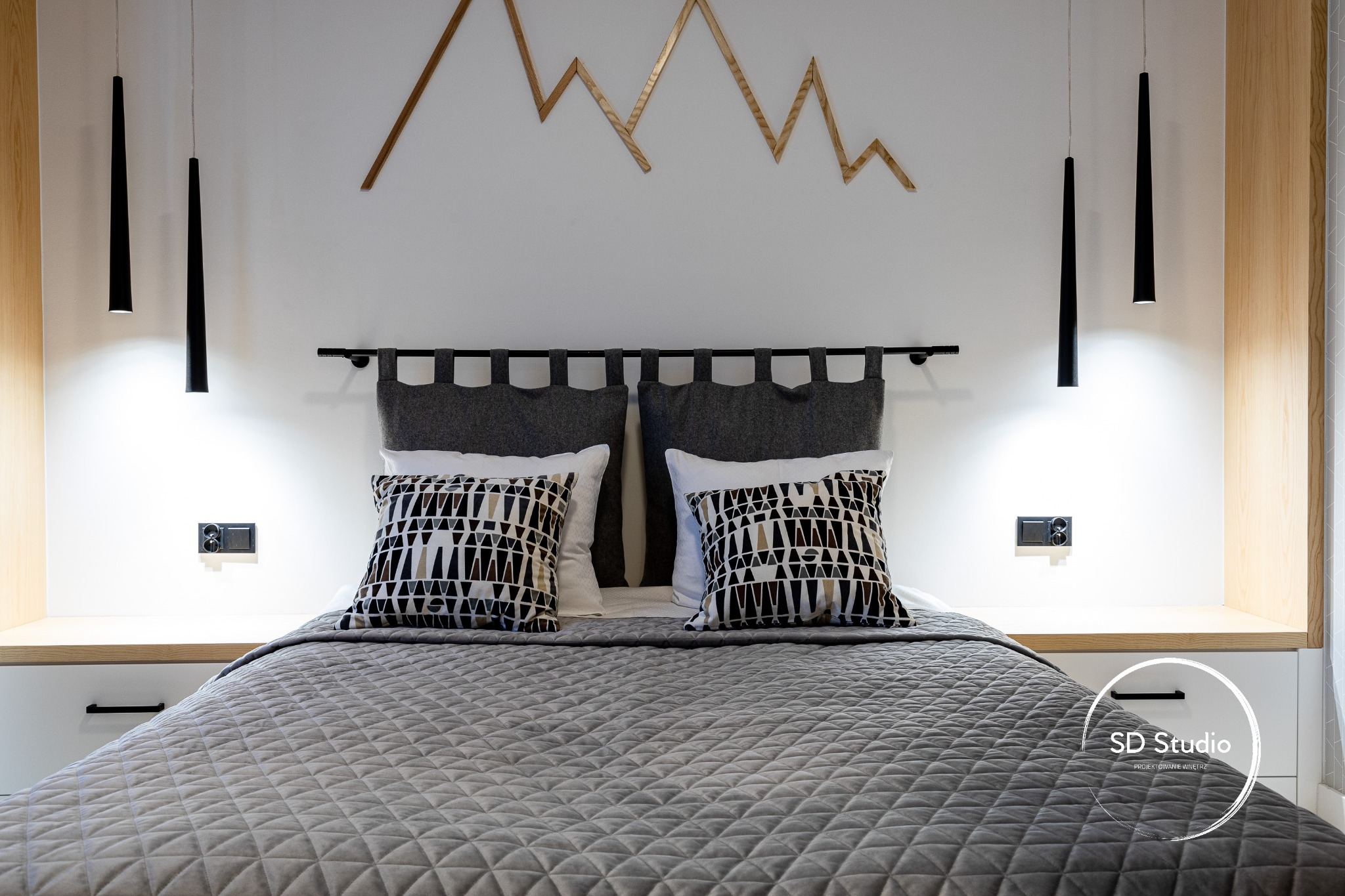 Nowoczesna sypialnia z szarą pikowaną narzutą, geometrycznymi poduszkami i drewnianą dekoracją ścienną w kształcie gór, oświetlona wiszącymi lampami tubami.