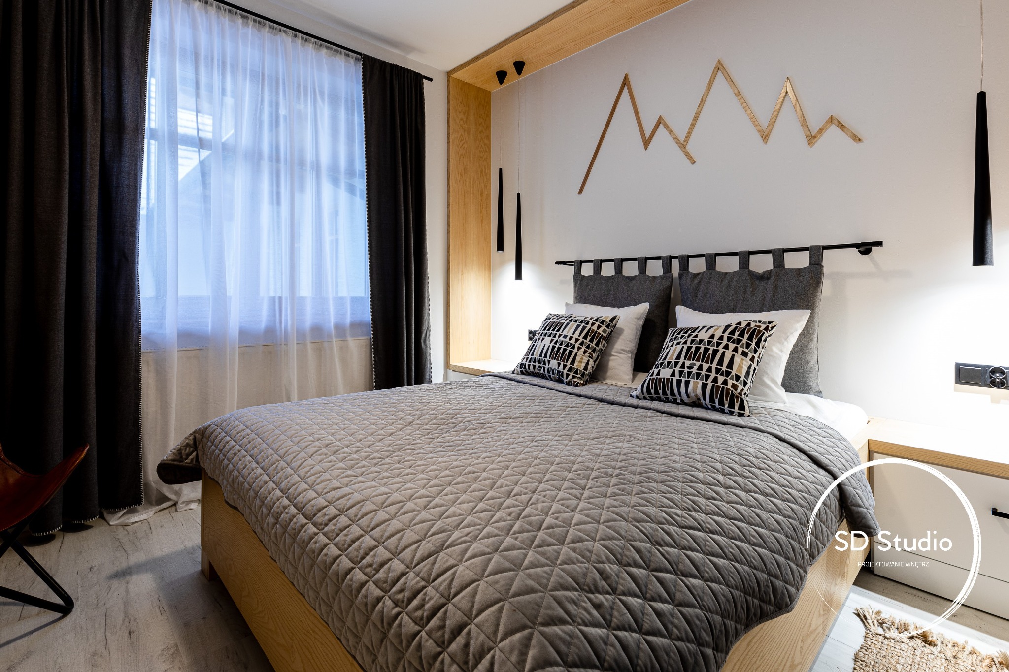 Nowoczesna sypialnia z łóżkiem na drewnianej ramie, szarą pikowaną narzutą, wzorzystymi poduszkami, drewnianą dekoracją ścienną w kształcie gór i wiszącymi lampami tubami.