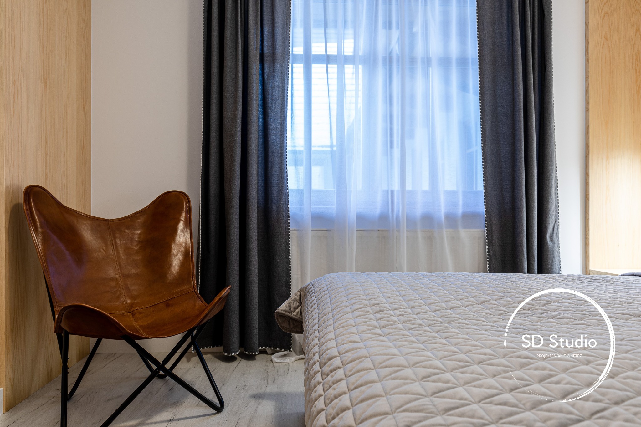 Sypialnia z łóżkiem w beżowej narzucie z geometrycznym wzorem, skórzanym fotelem typu motyl, szarymi zasłonami i drewnianą okładziną ściany.