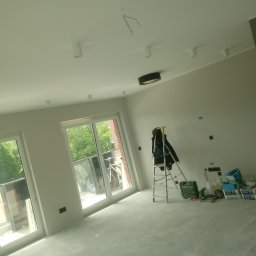 Łukasz Bąk - Wnętrze w trakcie remontu: świeżo pomalowane ściany, sufit z zamontowanymi lampami, drabina z kurtką, worki z zaprawą i narzędzia na podłodze przy oknie balkonowym.