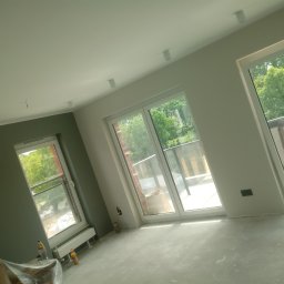 Łukasz Bąk - Wnętrze pokoju w trakcie remontu, widoczne trzy okna balkonowe, ściany częściowo pomalowane na szaro, podłoga przygotowana do dalszych prac wykończeniowych, widoczne butelki z napojami i zakryte...