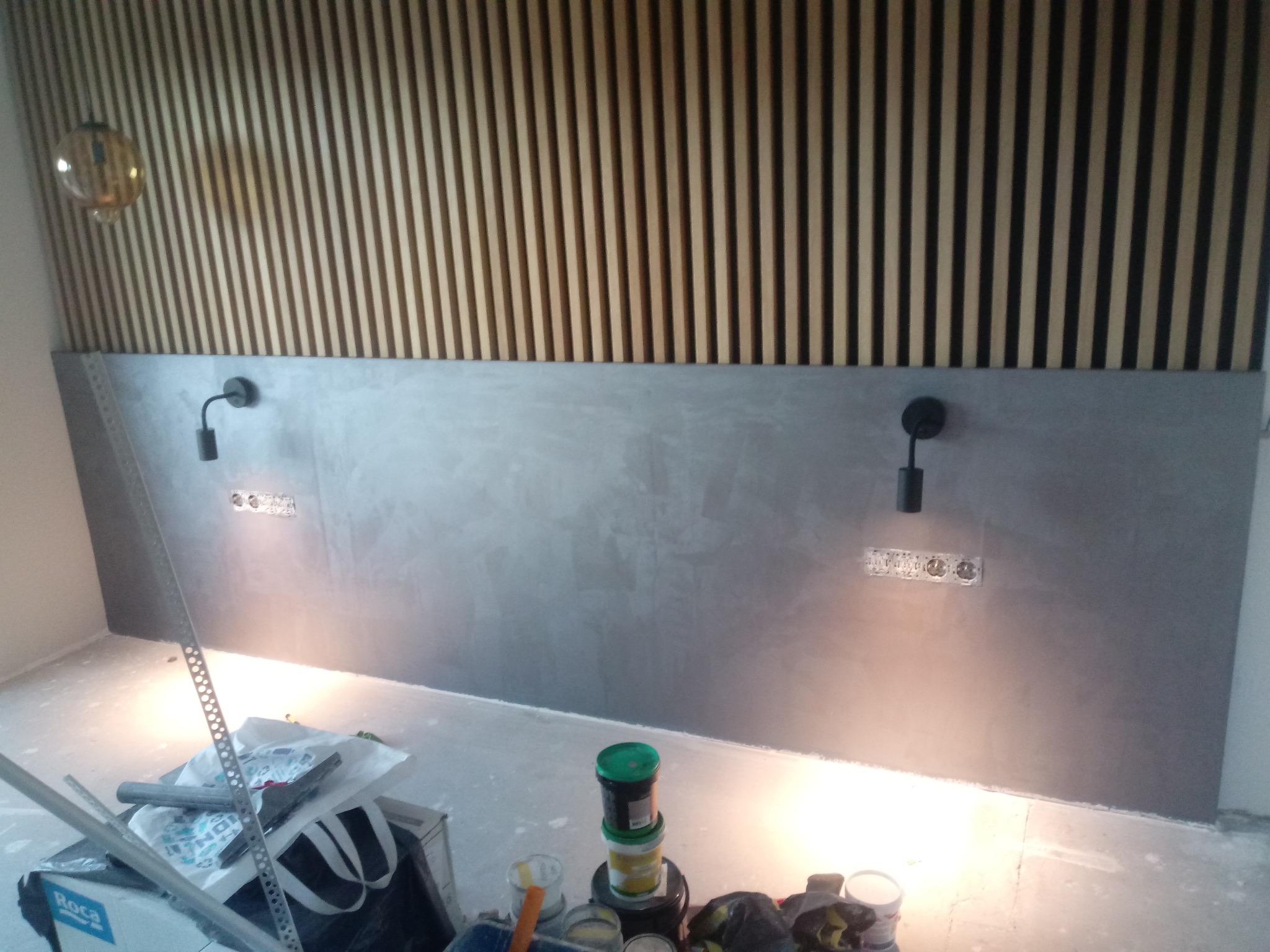 Ściana z szarą, gładką powierzchnią pod drewnianą dekoracją z pionowych listew, dwa czarne kinkiety zamontowane na ścianie, widoczne puszki elektryczne, na podłodze narzędzia i puszki z farbami.