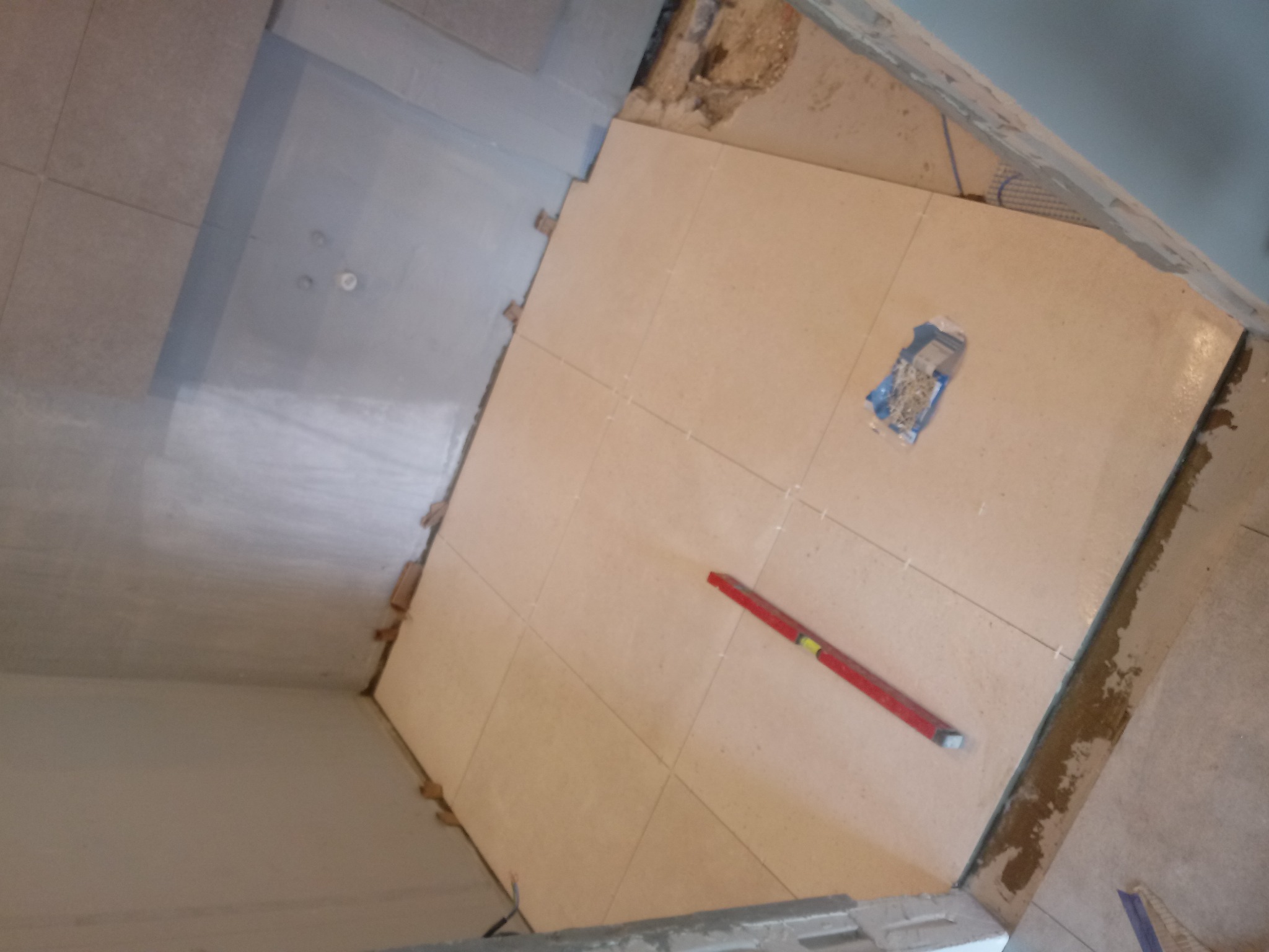 Ułożone beżowe płytki podłogowe w łazience, widoczna poziomica i opakowanie krzyżyków dystansowych, ściany przygotowane pod dalsze prace.